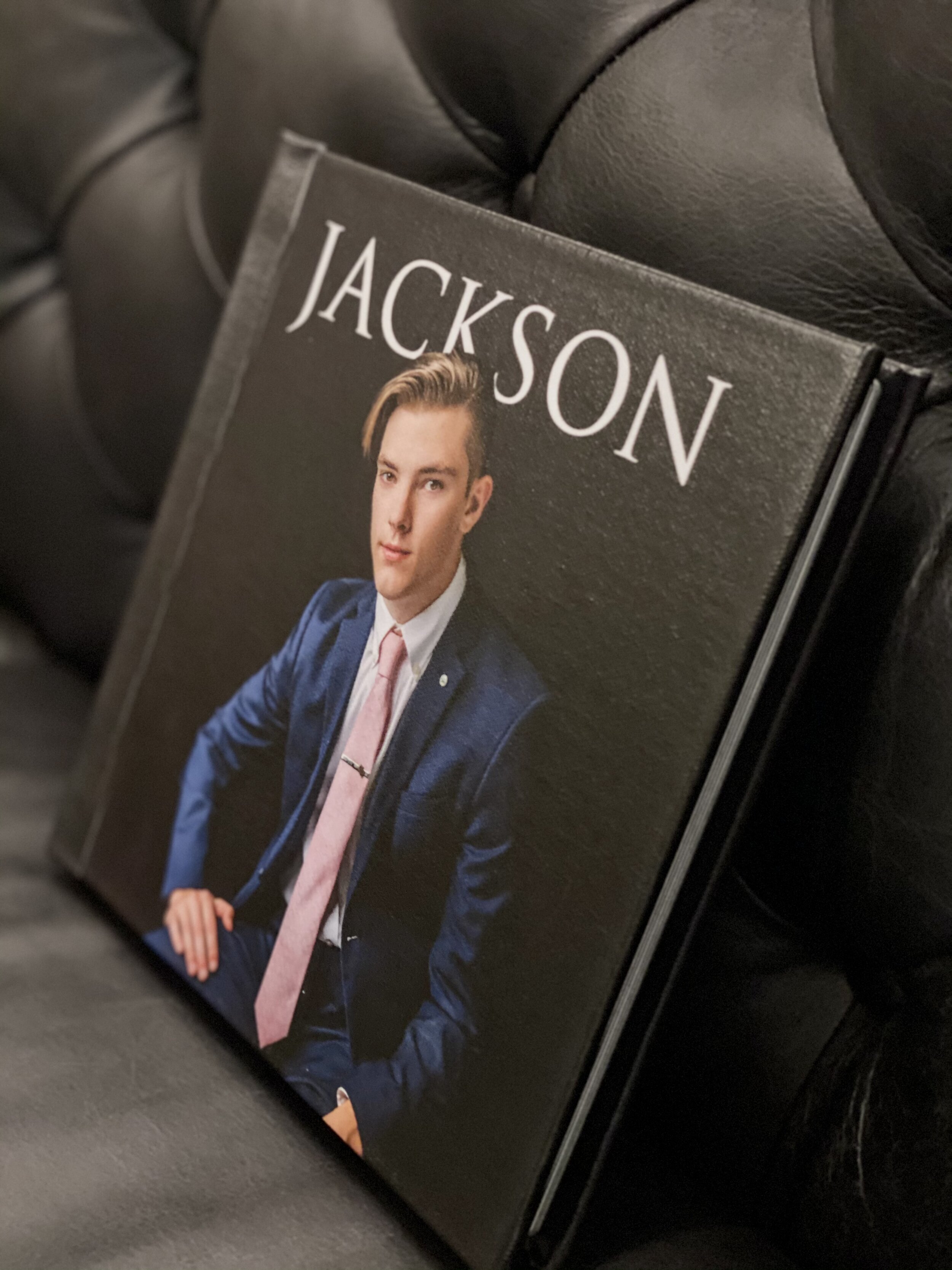 A book or magazine with the title 'JACKSON' and a portrait of a young man with styled blond hair in a blue suit and pink tie, positioned on a black leather surface.