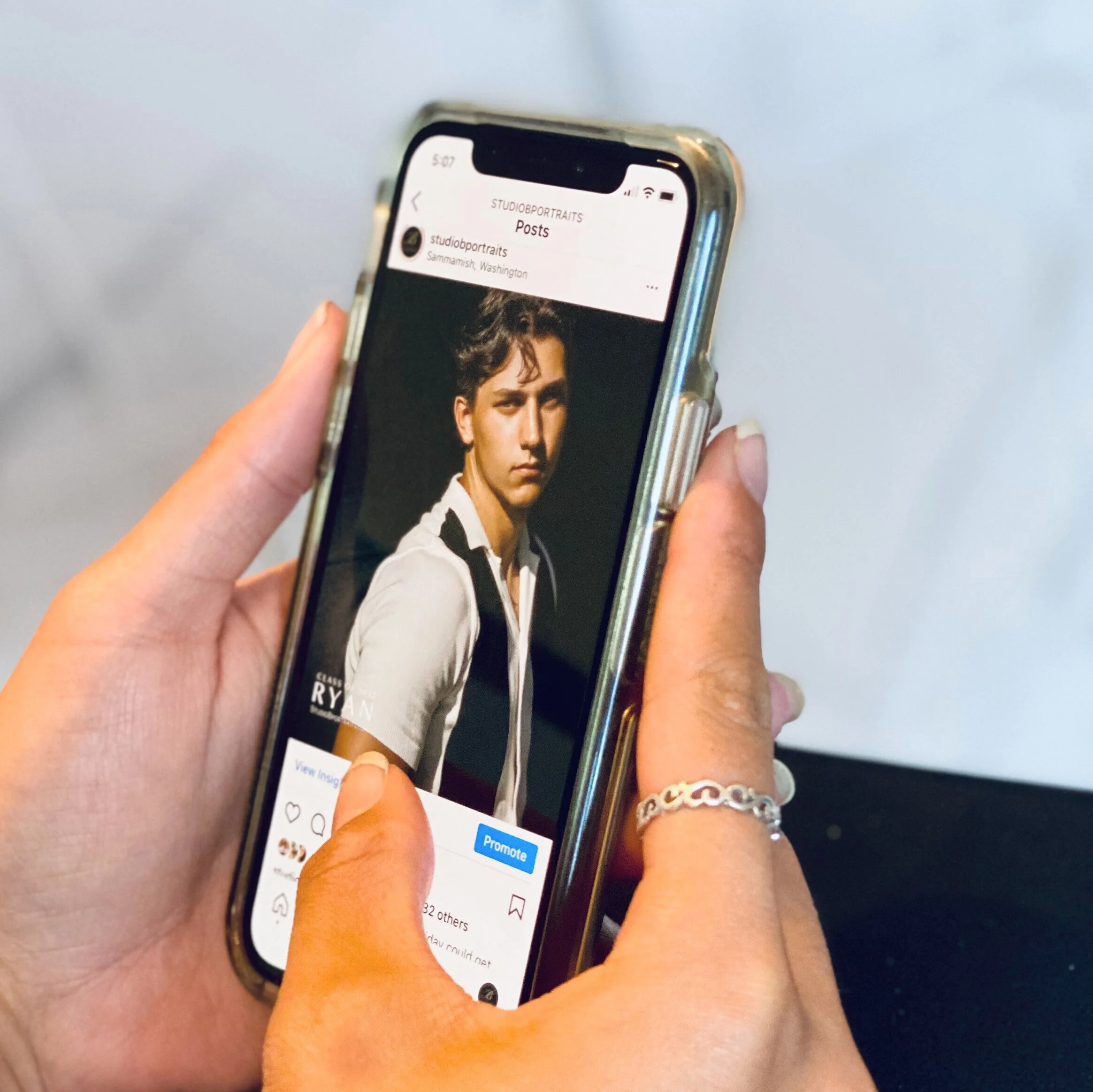Person holding a smartphone displaying an Instagram post of a young man with dark hair, wearing a white shirt and black tie.