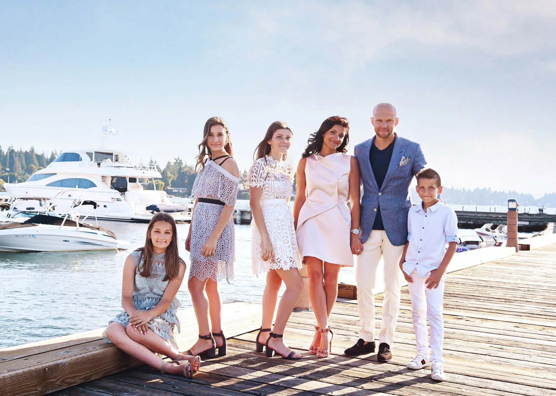 Family of seven dressed in stylish clothes standing on a wooden dock by the water with boats and yachts in the background, under a clear blue sky.