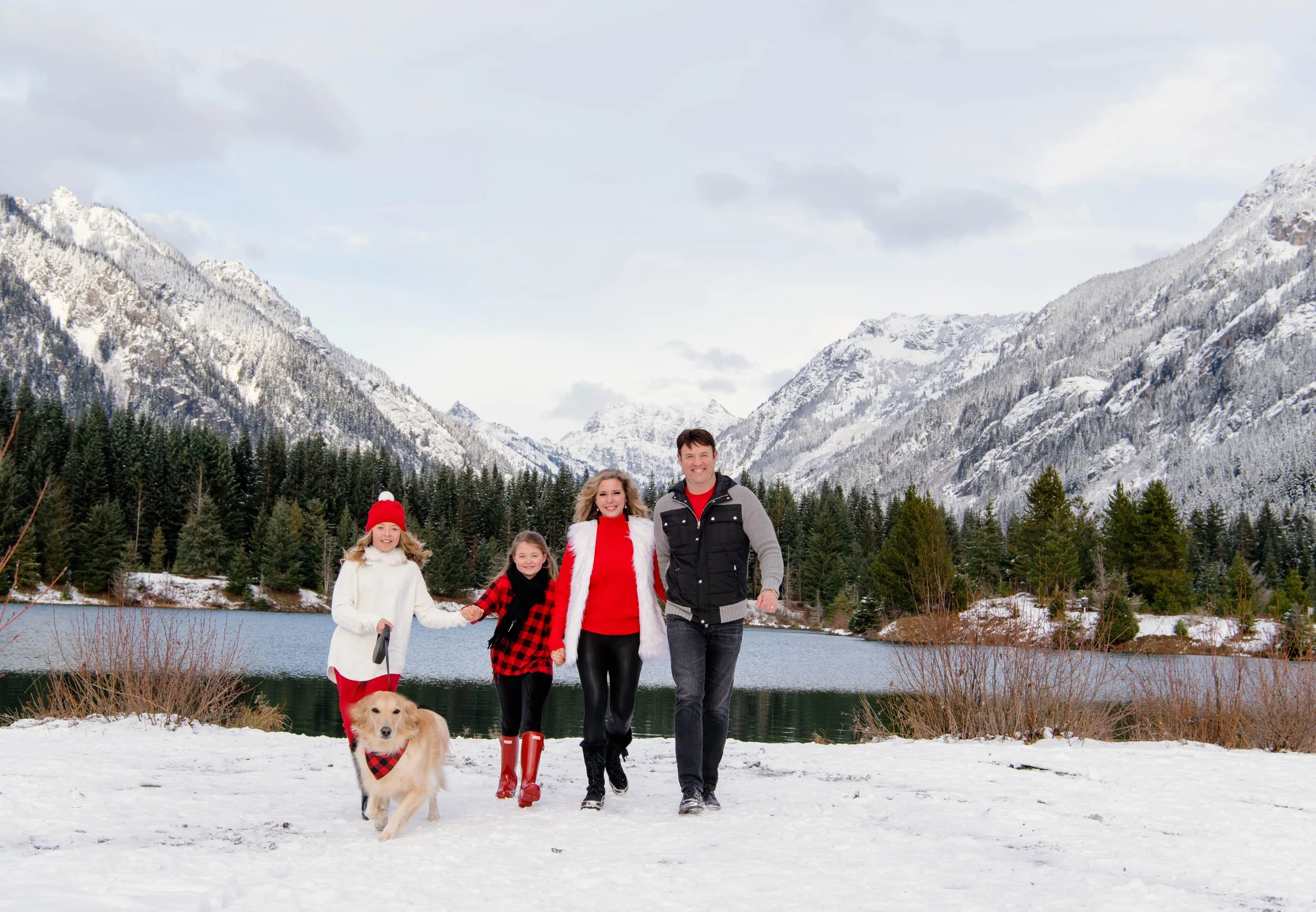 Family of four with a dog enjoying a snowy landscape with snow-covered mountains, a lake, and trees in the background.
