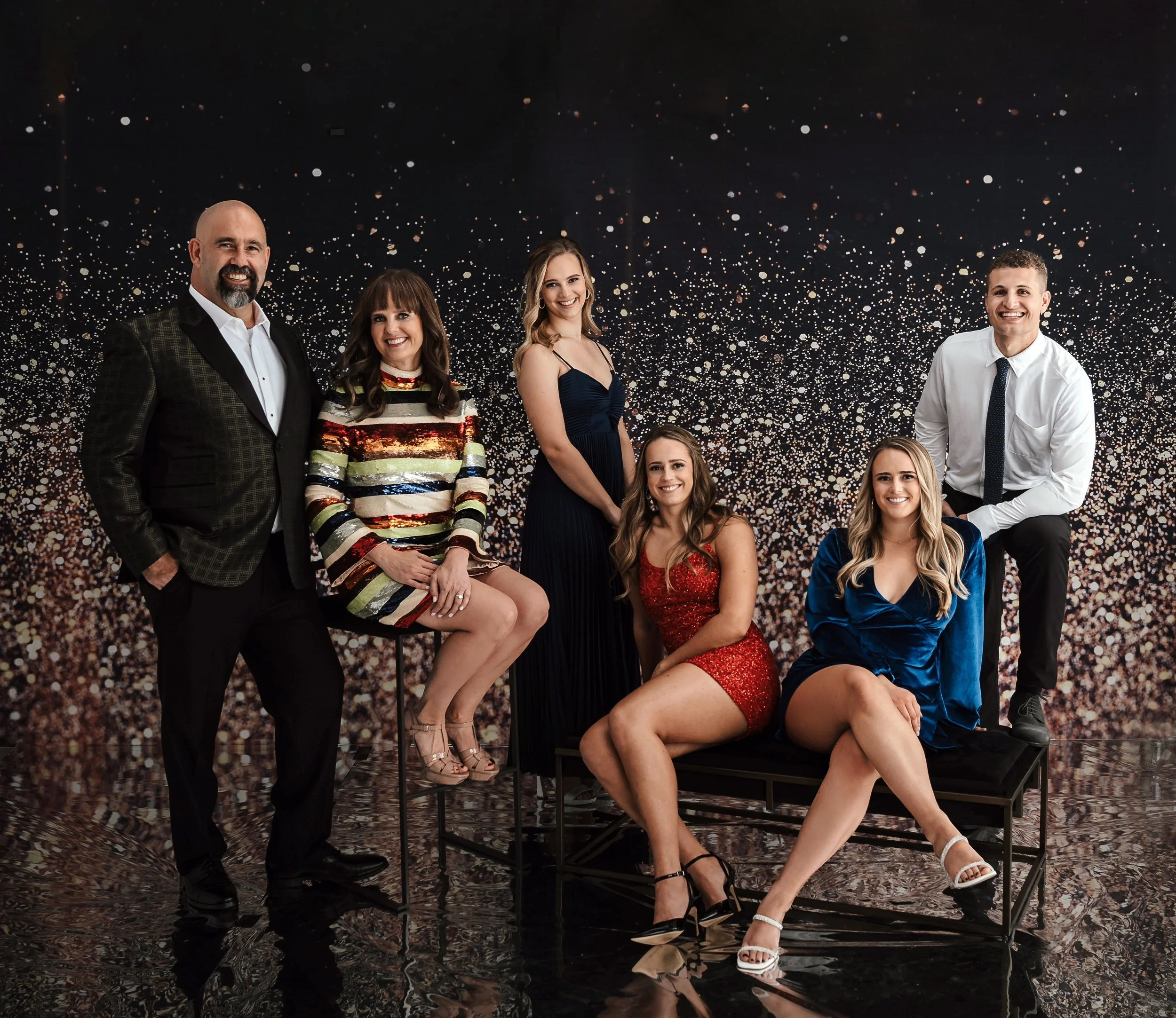 Group of seven people dressed in formal attire, posing together in front of a dark, sparkling backdrop for a photo.
