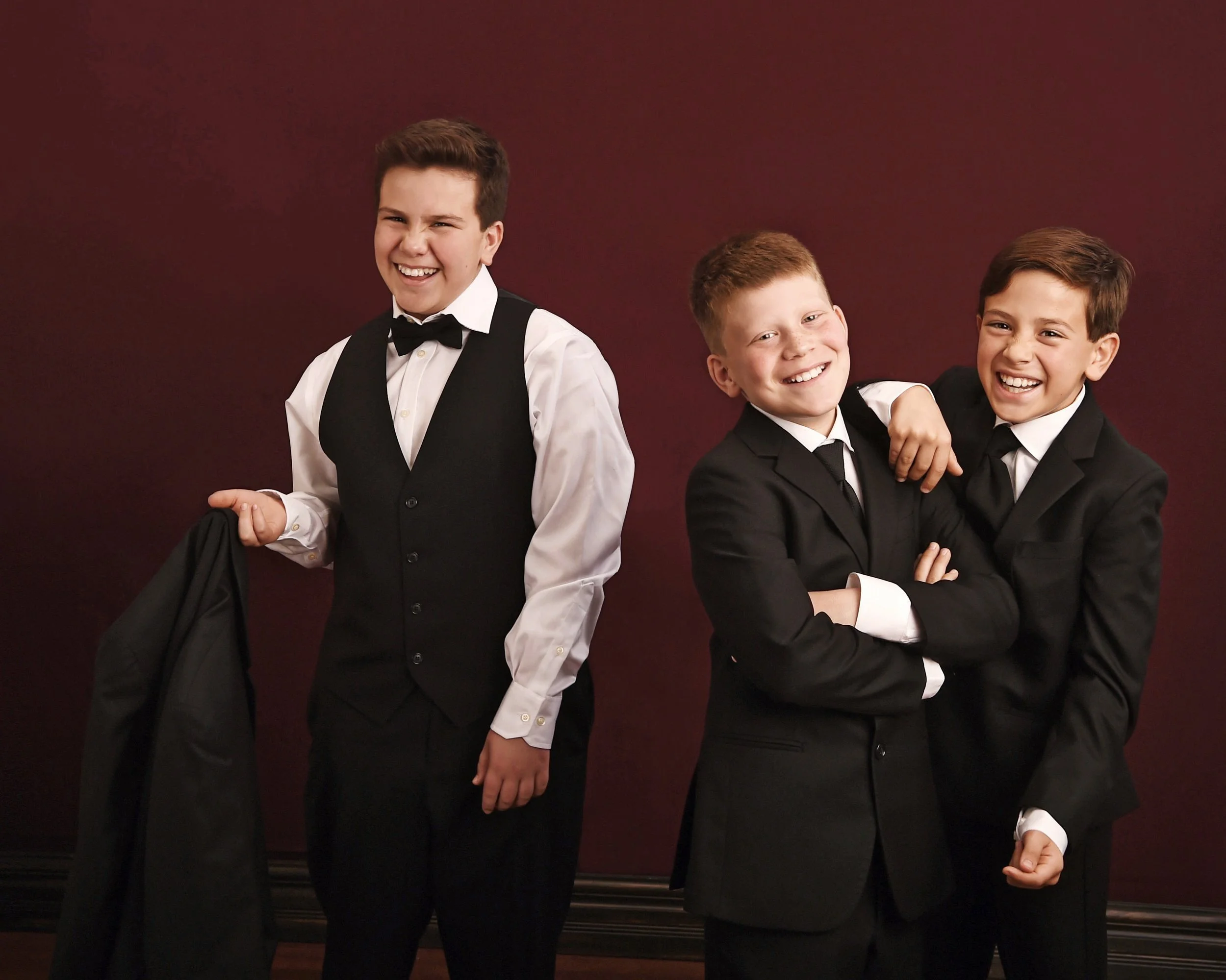 Three boys dressed in black tuxedos and white shirts smiling and laughing together against a dark red background.
