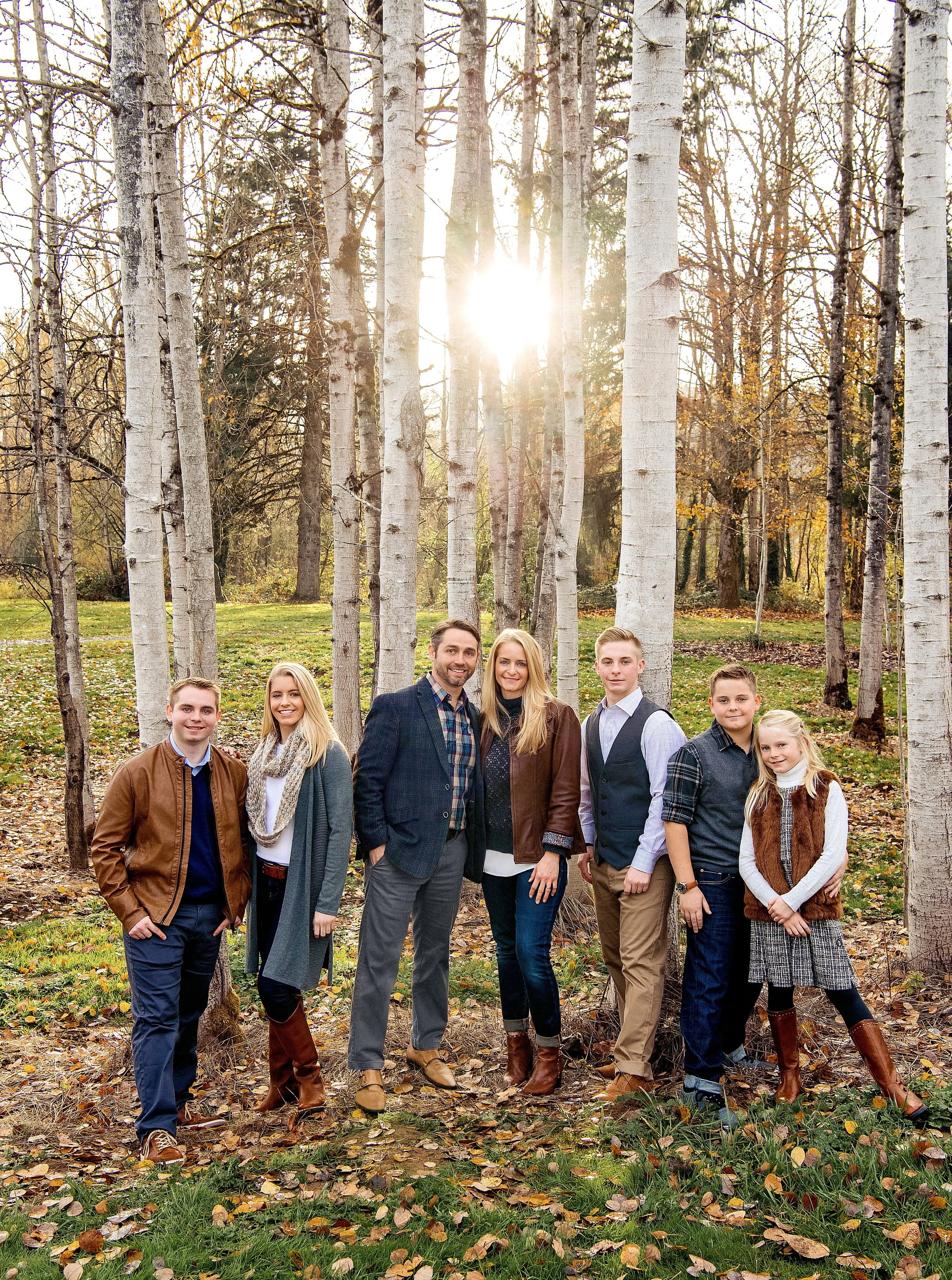 A family of seven standing outdoors among tall, leafless trees in a park during autumn, smiling at the camera with the sun shining through the trees behind them.