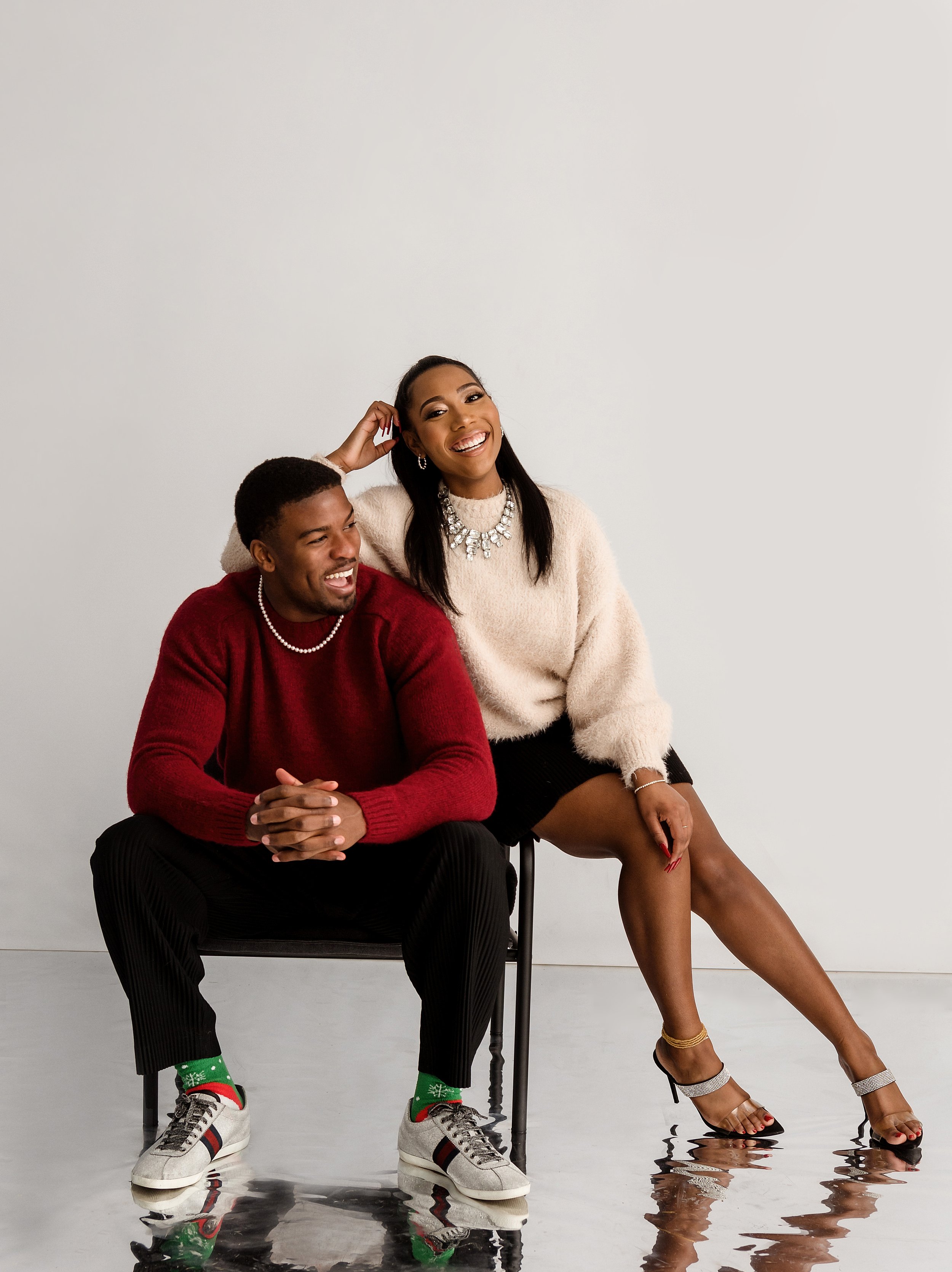A smiling man and woman sitting together, with the woman sitting on a chair and the man sitting on the floor, both dressed festively with jewelry and the woman wearing heels, against a plain white background.