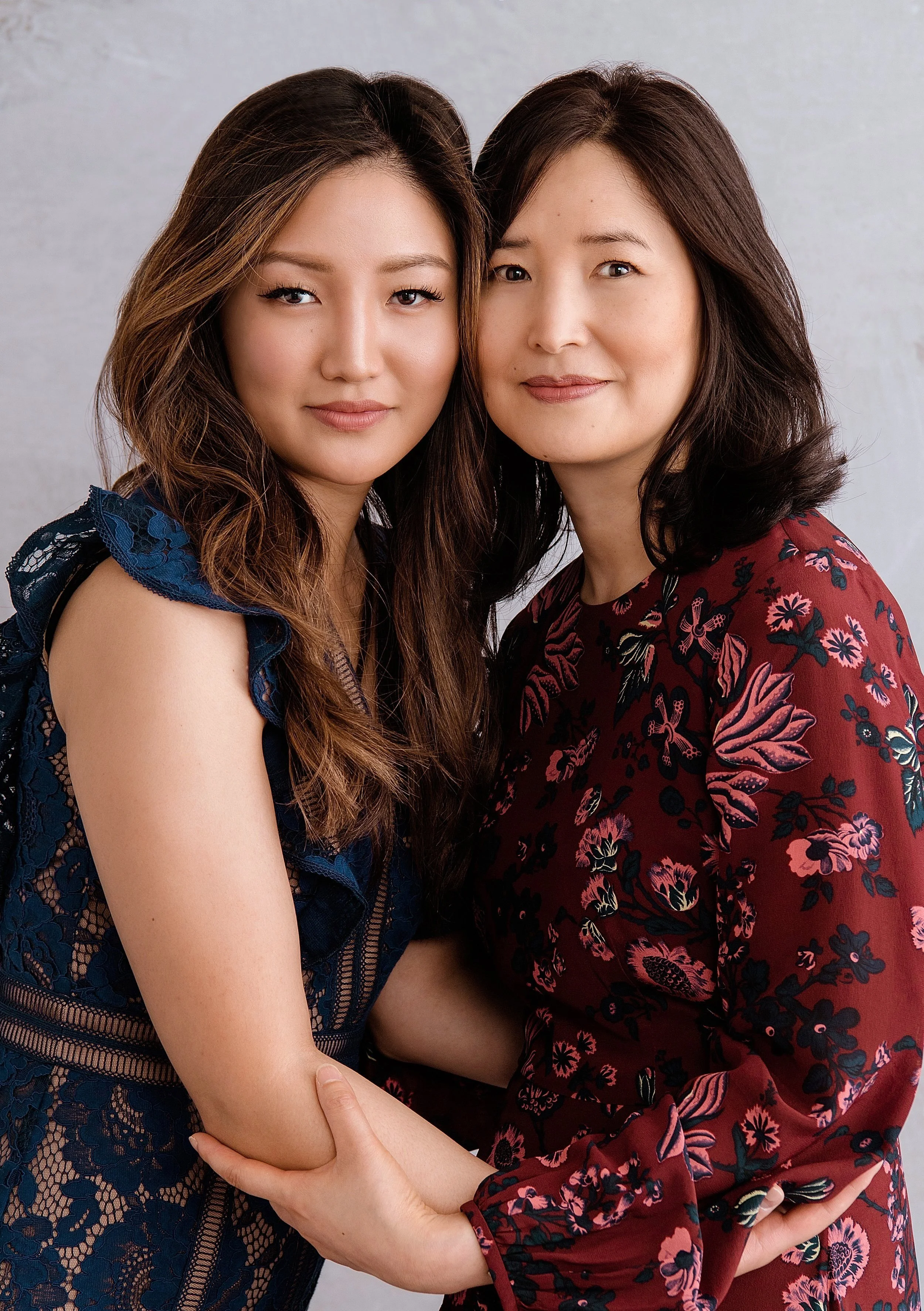 Two women embracing, one in a blue lace dress, the other in a red floral dress, smiling at the camera.