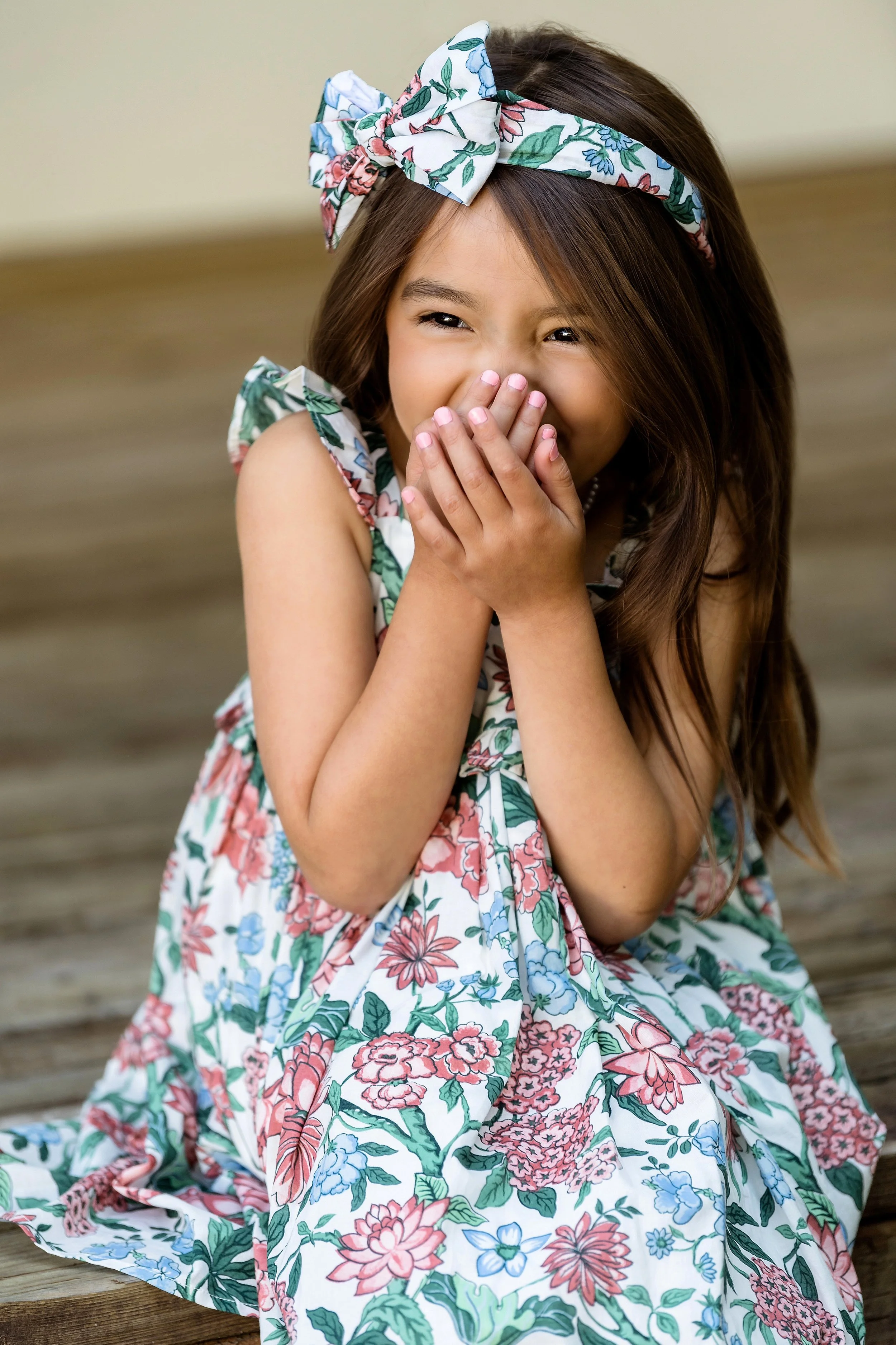 A young girl with long brown hair, wearing a floral dress and headband, is sitting on a wooden surface and laughing with her hand covering her mouth.