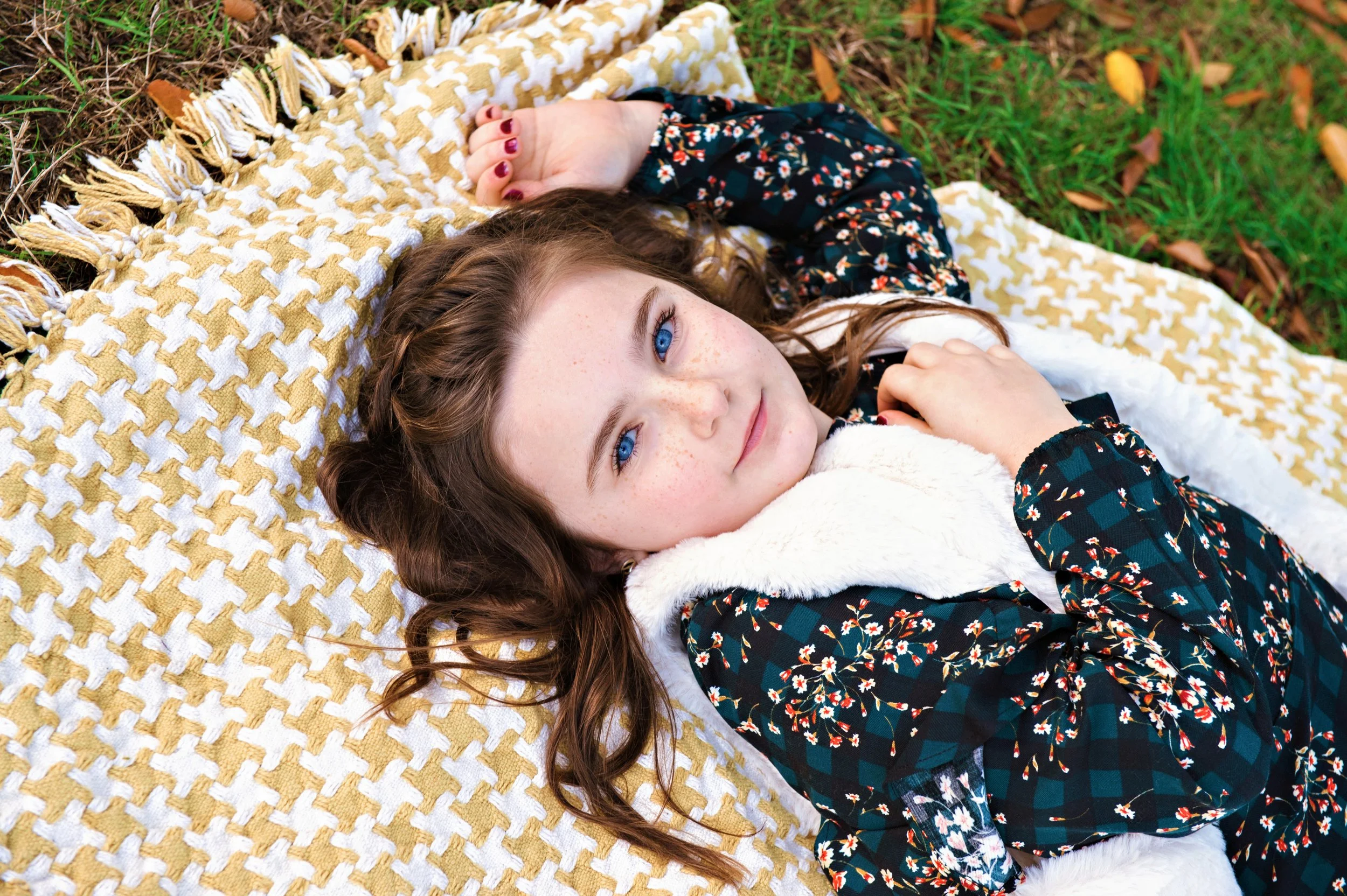 Young girl lying on a yellow and white checkered blanket outdoors on grass, with brown curly hair, blue eyes, and freckles, wearing a dark floral jacket with a white furry collar.