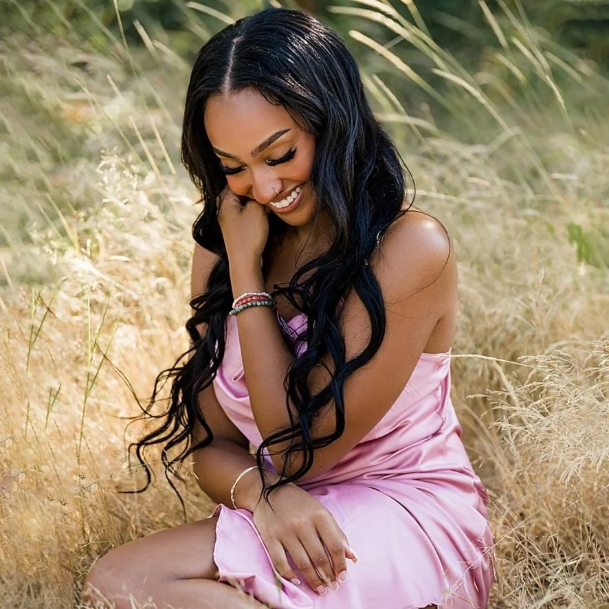 A woman with long black hair sitting in tall, golden grass, wearing a pink satin dress and smiling joyfully.