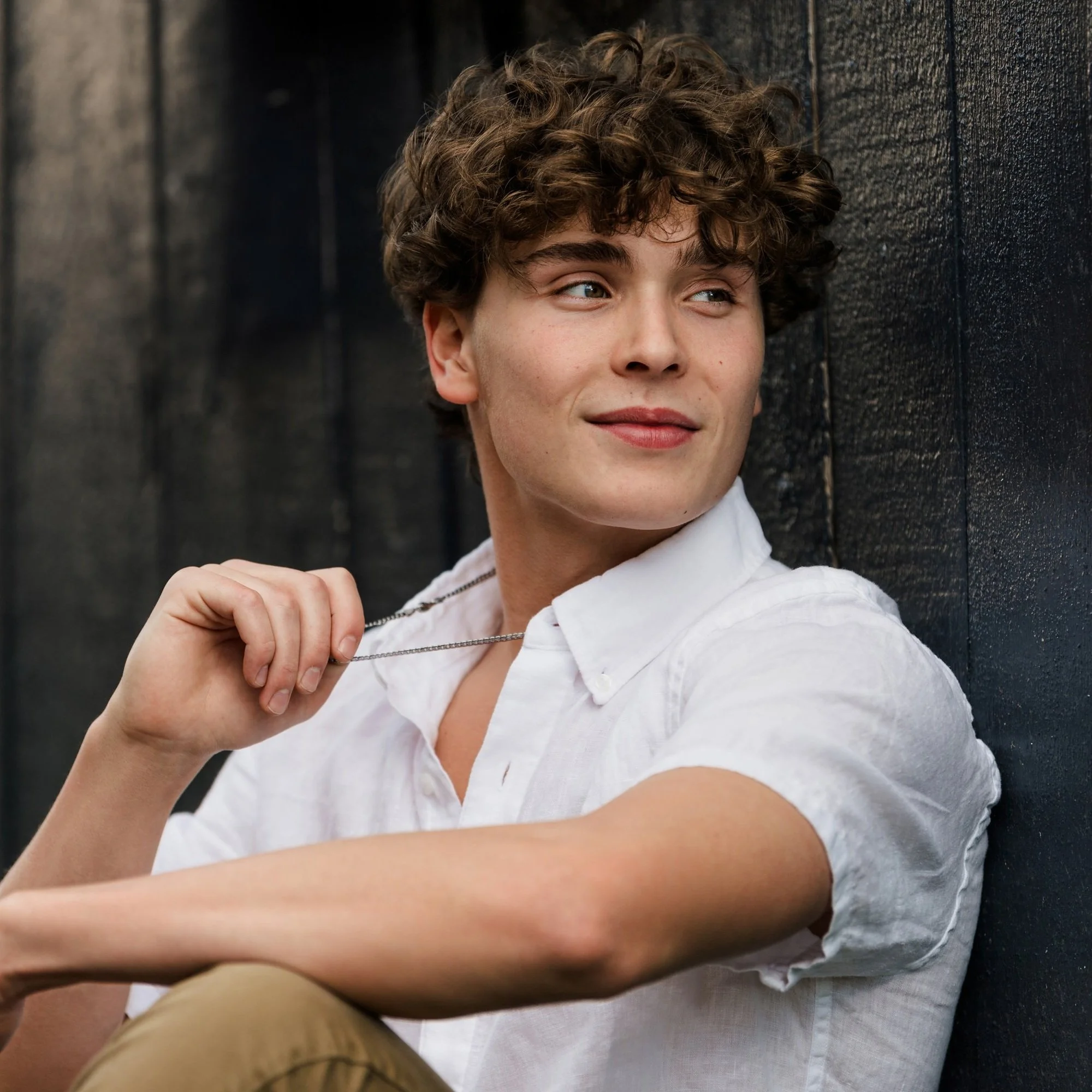 A young man with curly brown hair wearing a white short-sleeve shirt, sitting against a dark wooden wall, holding a necklace and looking to his right with a slight smile.