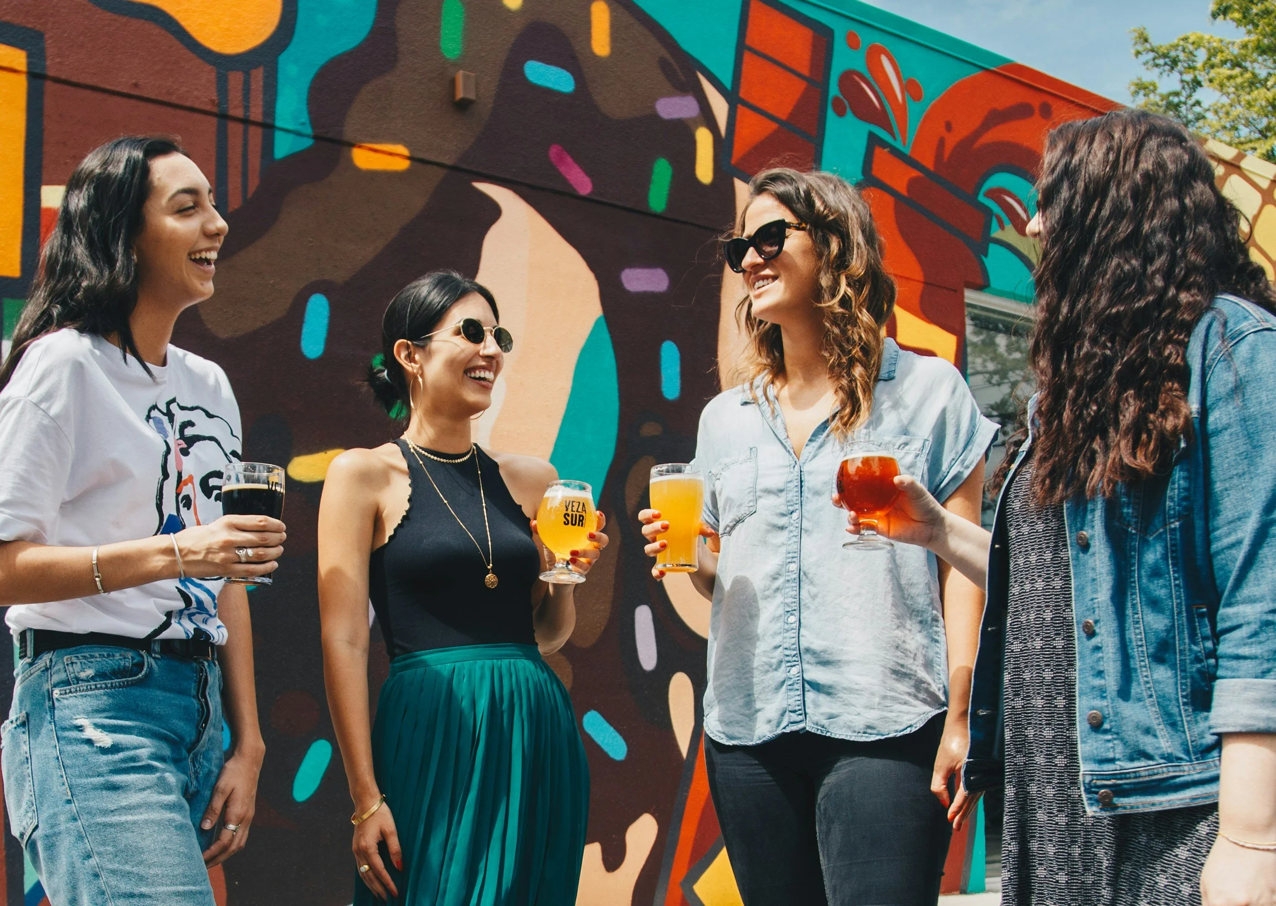 Five women sharing drinks and laughing outdoors in front of a colorful mural.