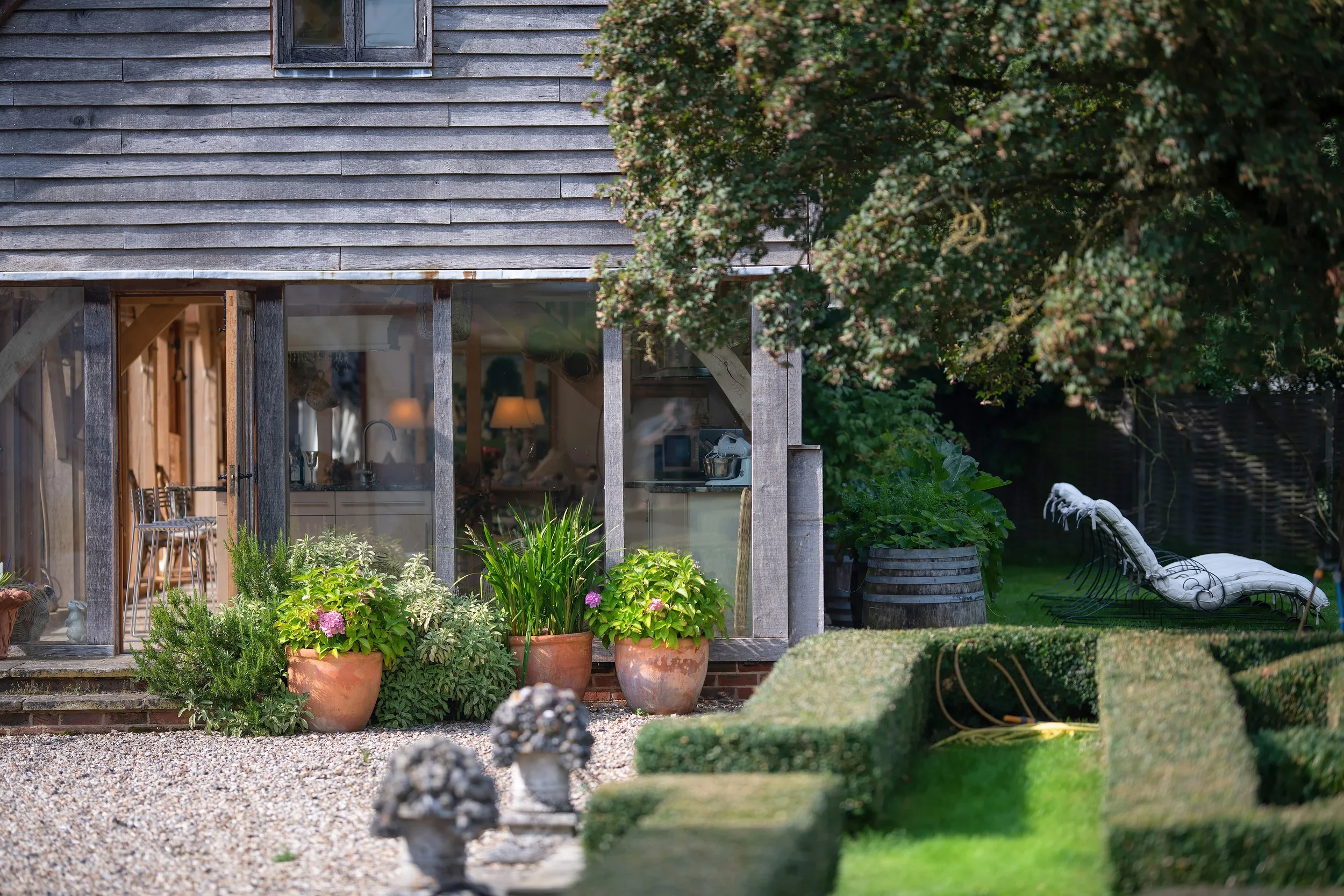 A view of a house with a screened porch, potted plants, and outdoor seating, surrounded by greenery and a lawn.