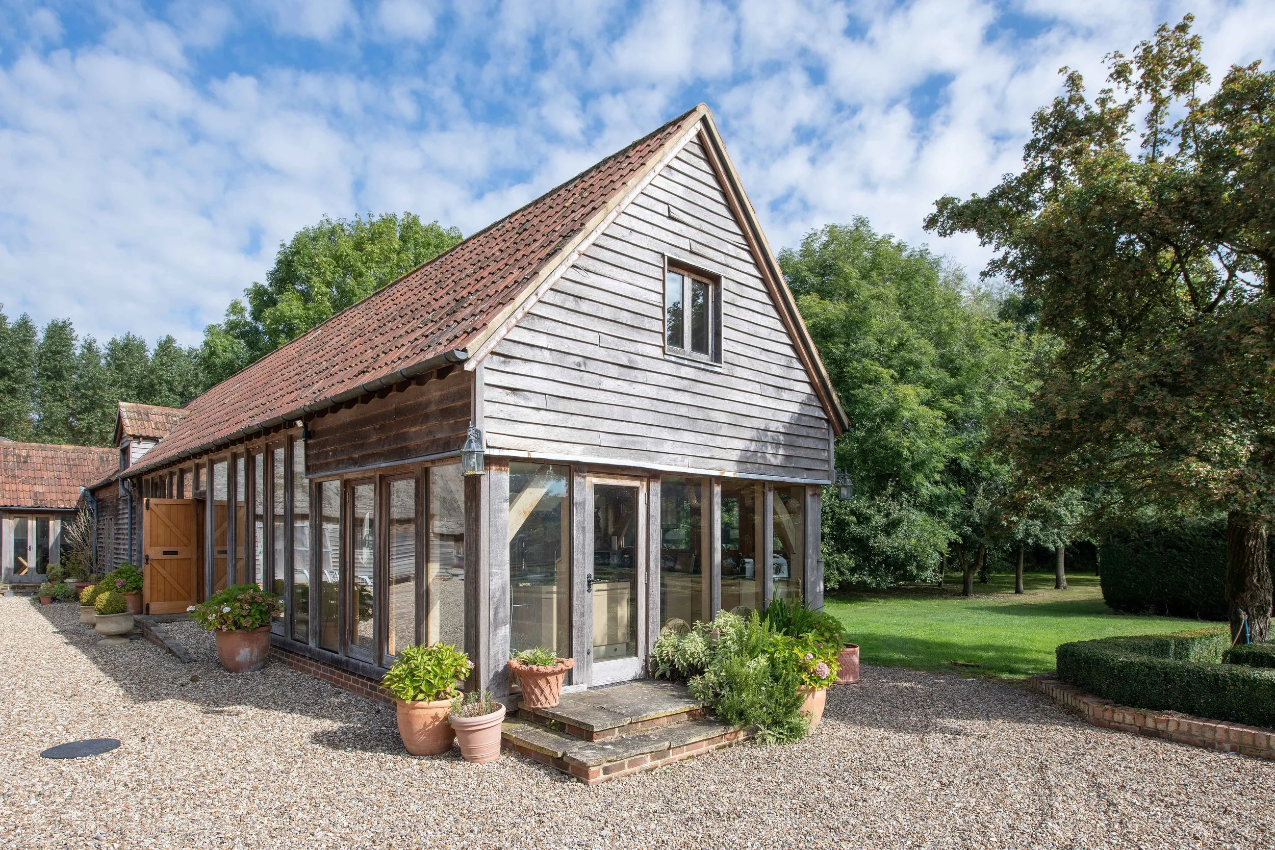 A rustic house with a wooden exterior, large glass windows, potted plants, and a gravel pathway, surrounded by green trees and a lawn.