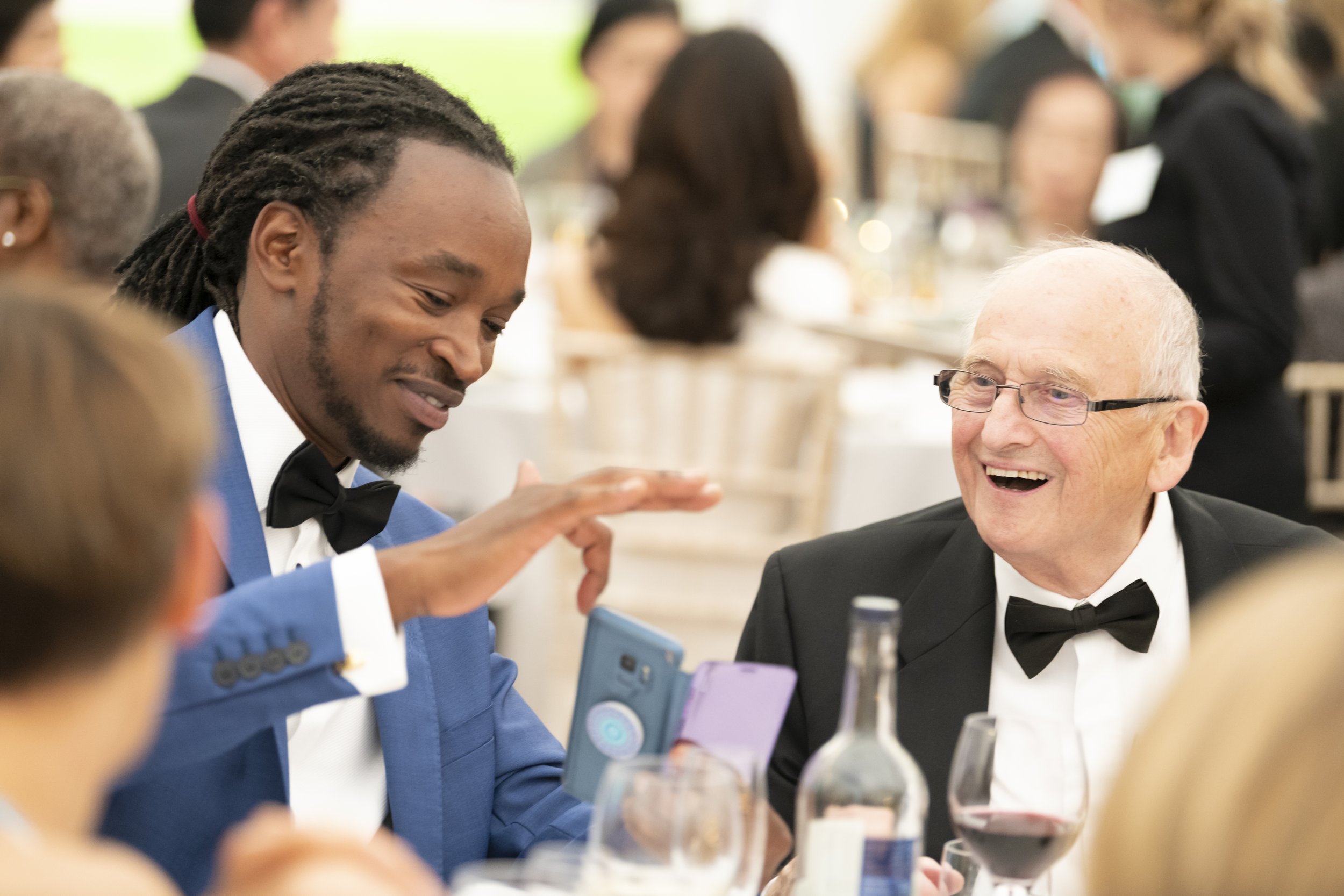 Two men in tuxedos at a formal event, one with dreadlocks and the other with glasses, smiling and looking at a smartphone.