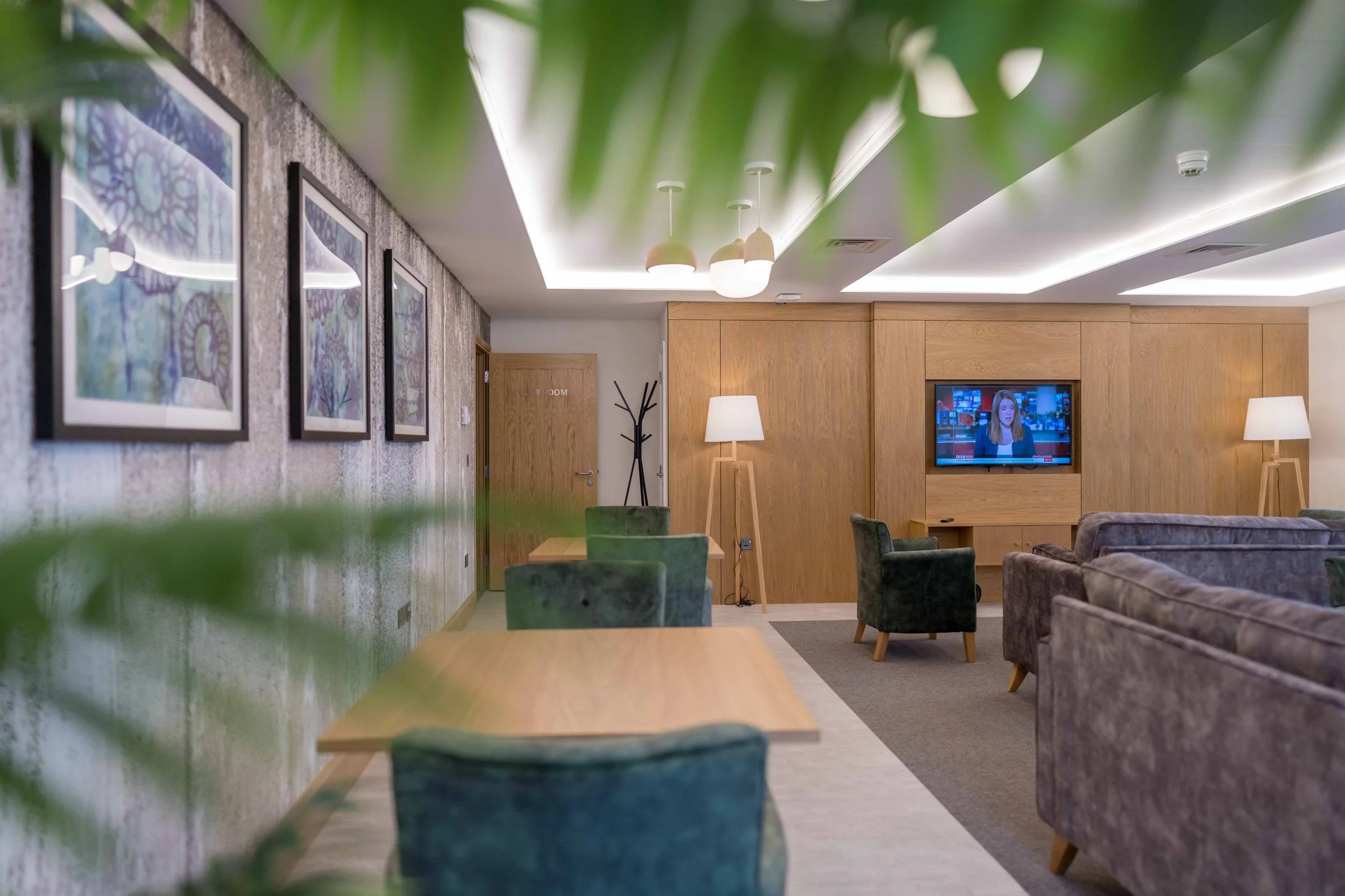 A cozy lounge or waiting area with wooden wall paneling, a TV mounted on the wall, and multiple sitting chairs and couches. The room has modern lighting and framed artwork on textured walls, with green plants partially visible in the foreground.