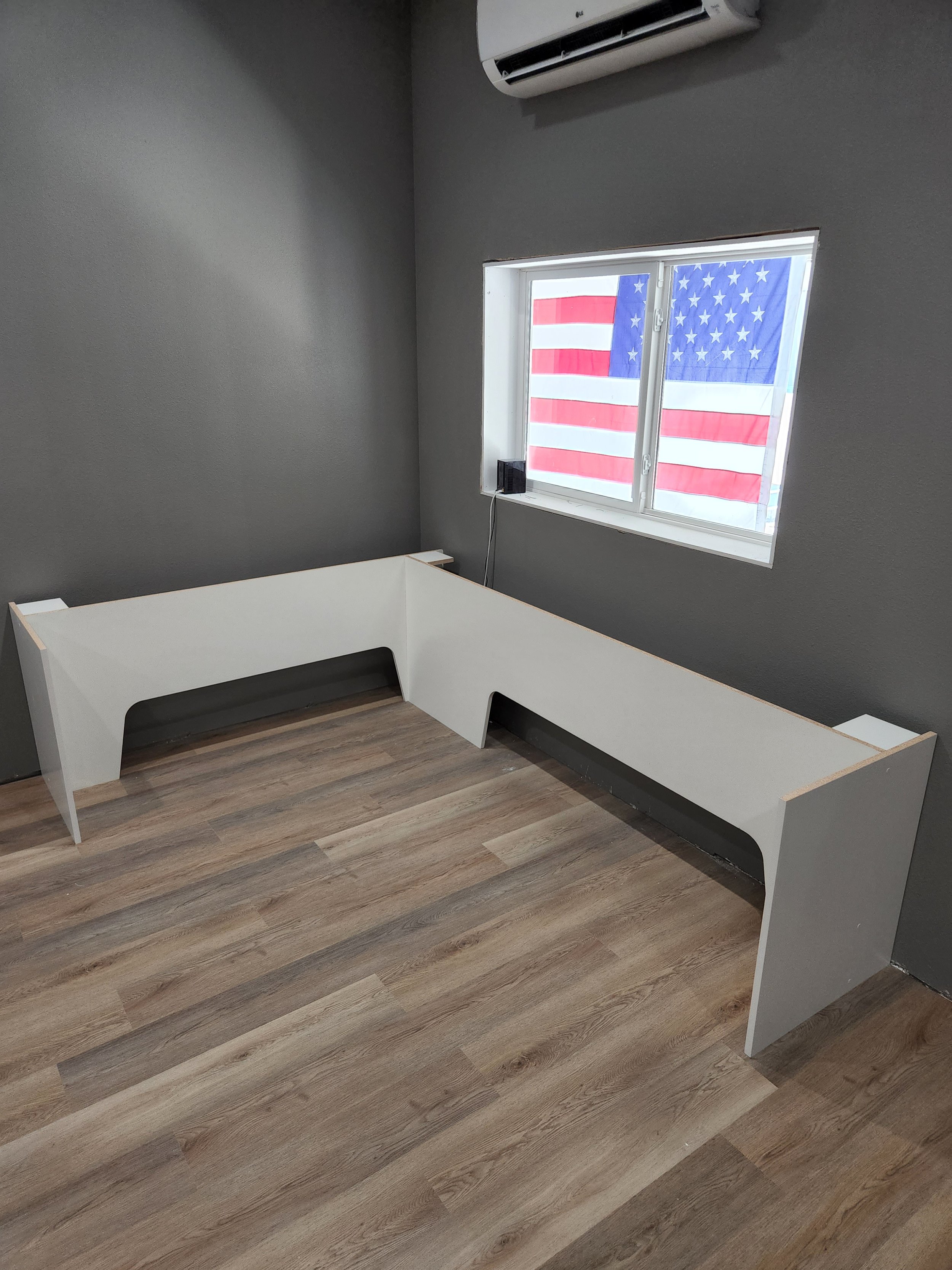 Empty room with gray walls, a white L-shaped desk, a window with an American flag, and an air conditioning unit.