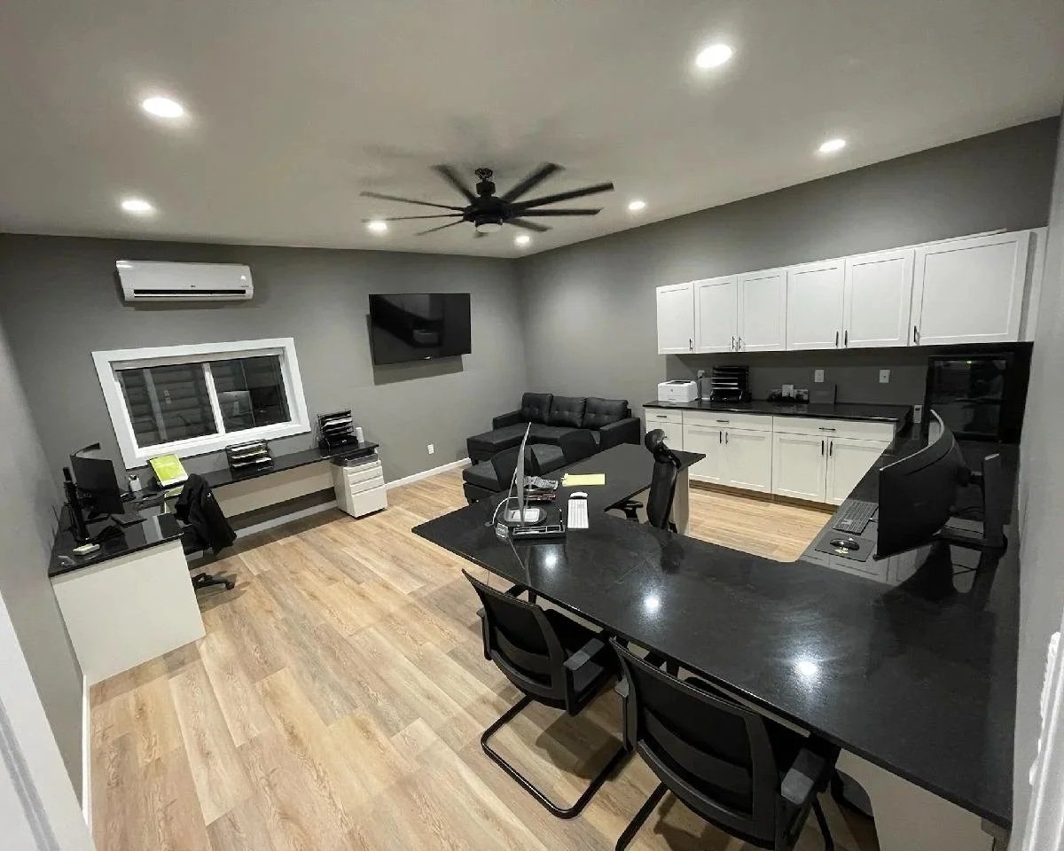Modern office and lounge space with black desks, computers, a black sectional sofa, a wall-mounted TV, and white cabinets on gray walls, illuminated by recessed ceiling lights.