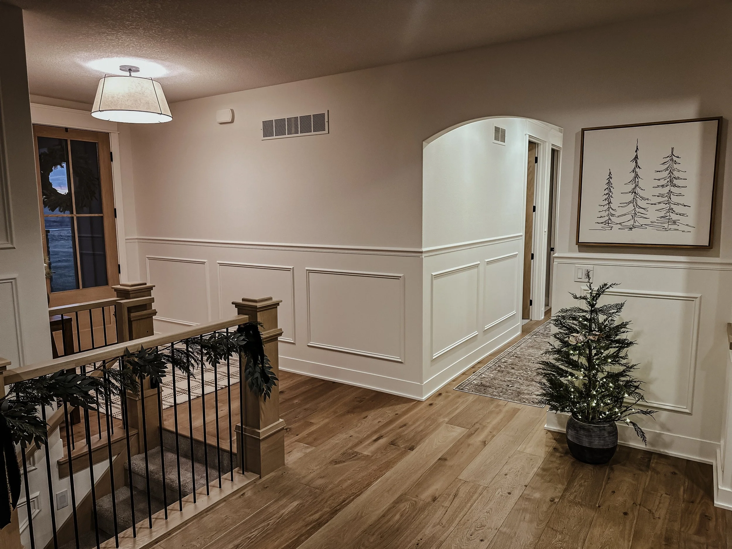 Interior of a home with hardwood floors, white walls with decorative molding, a staircase with wooden and black metal railing, a potted small Christmas tree, and framed artwork of trees on the wall.