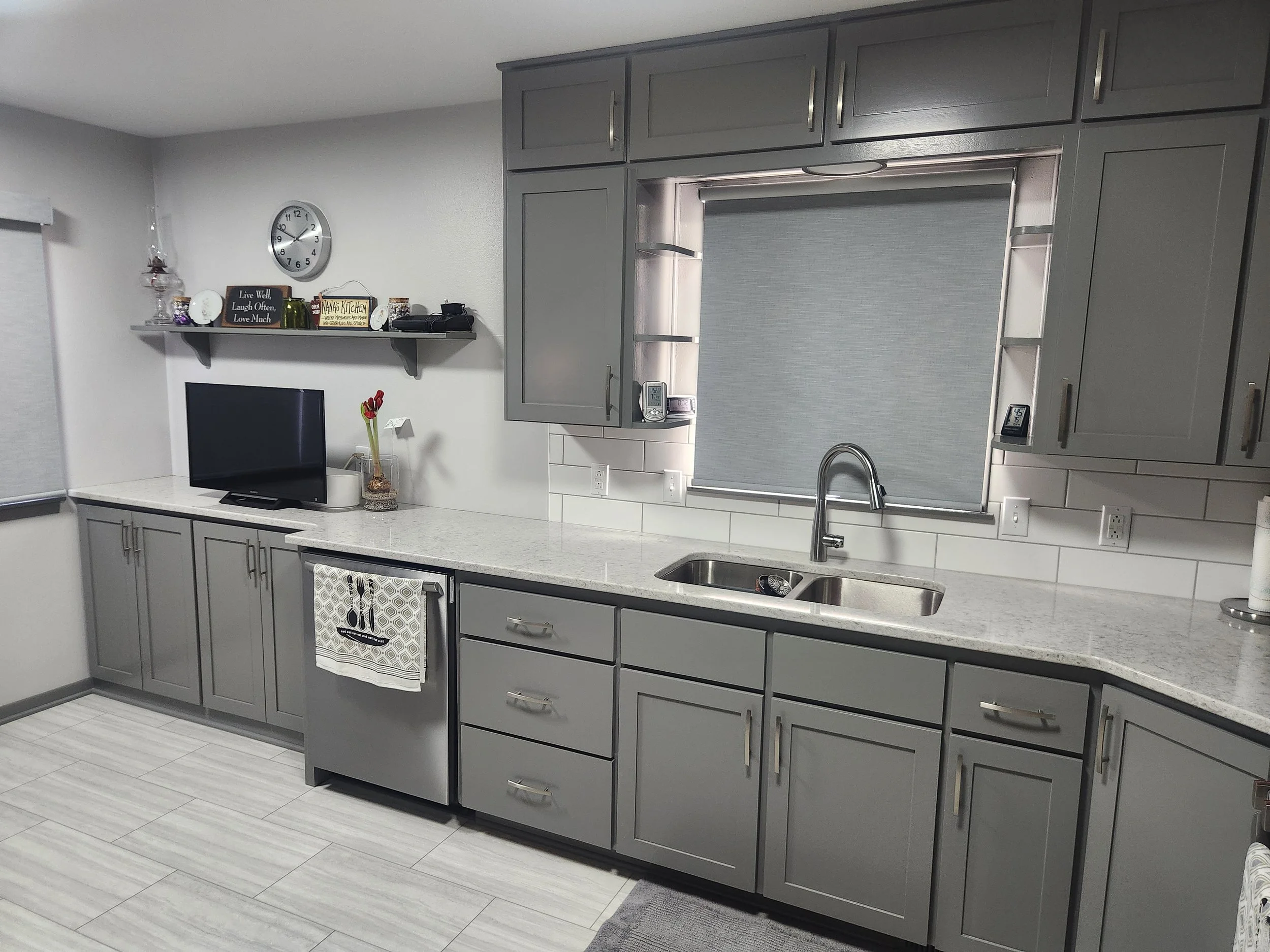Gray kitchen cabinets with a marble countertop, a double sink, and a window with a gray roller shade.