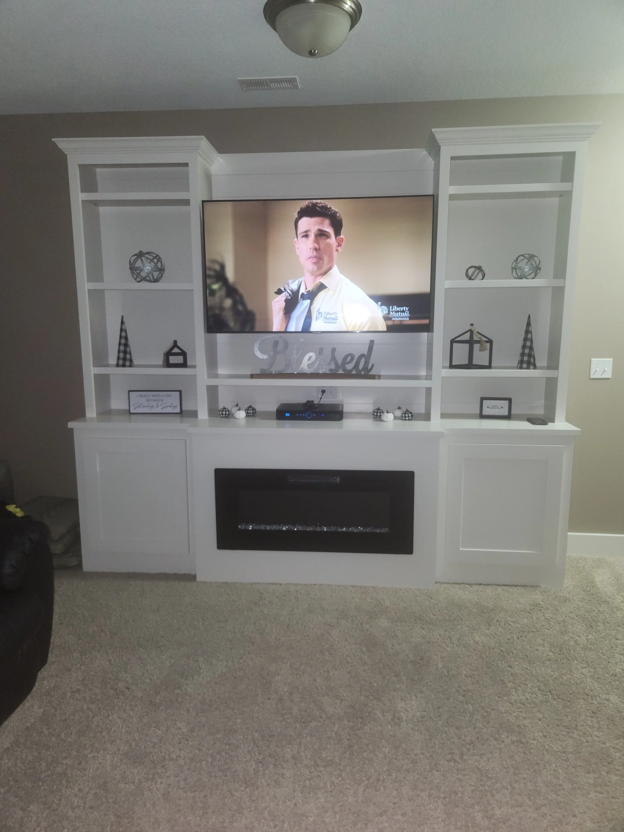 White entertainment center with shelves, a TV showing a man in a white shirt, and a fireplace below the TV in a living room.