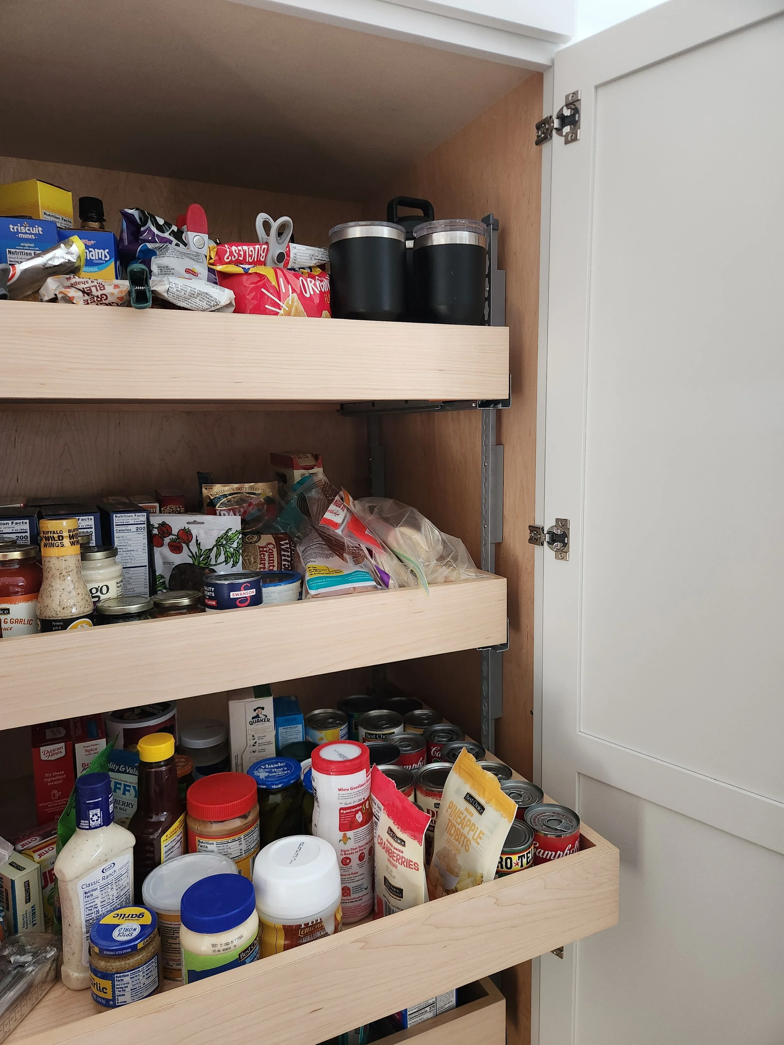 Open kitchen pantry with three wooden shelves filled with snacks, canned goods, bottles, and jars.