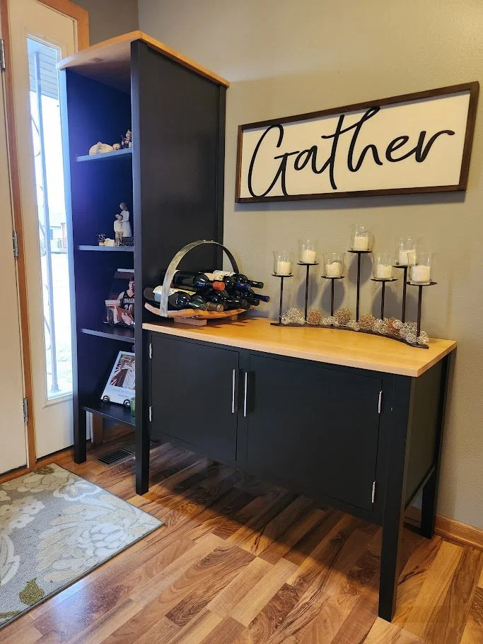 A black and wooden sideboard with a wine rack, decorative candles, and a "Gather" sign on the wall above it, near a door with a window.