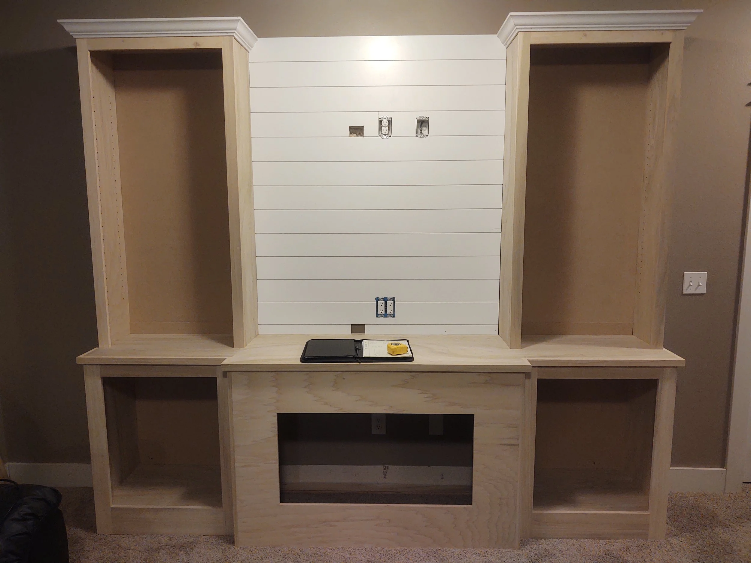Wooden built-in shelving unit with two open compartments on each side, a small central opening, and a flat top surface, against a wall with white horizontal paneling, with electrical outlets and tools on top.
