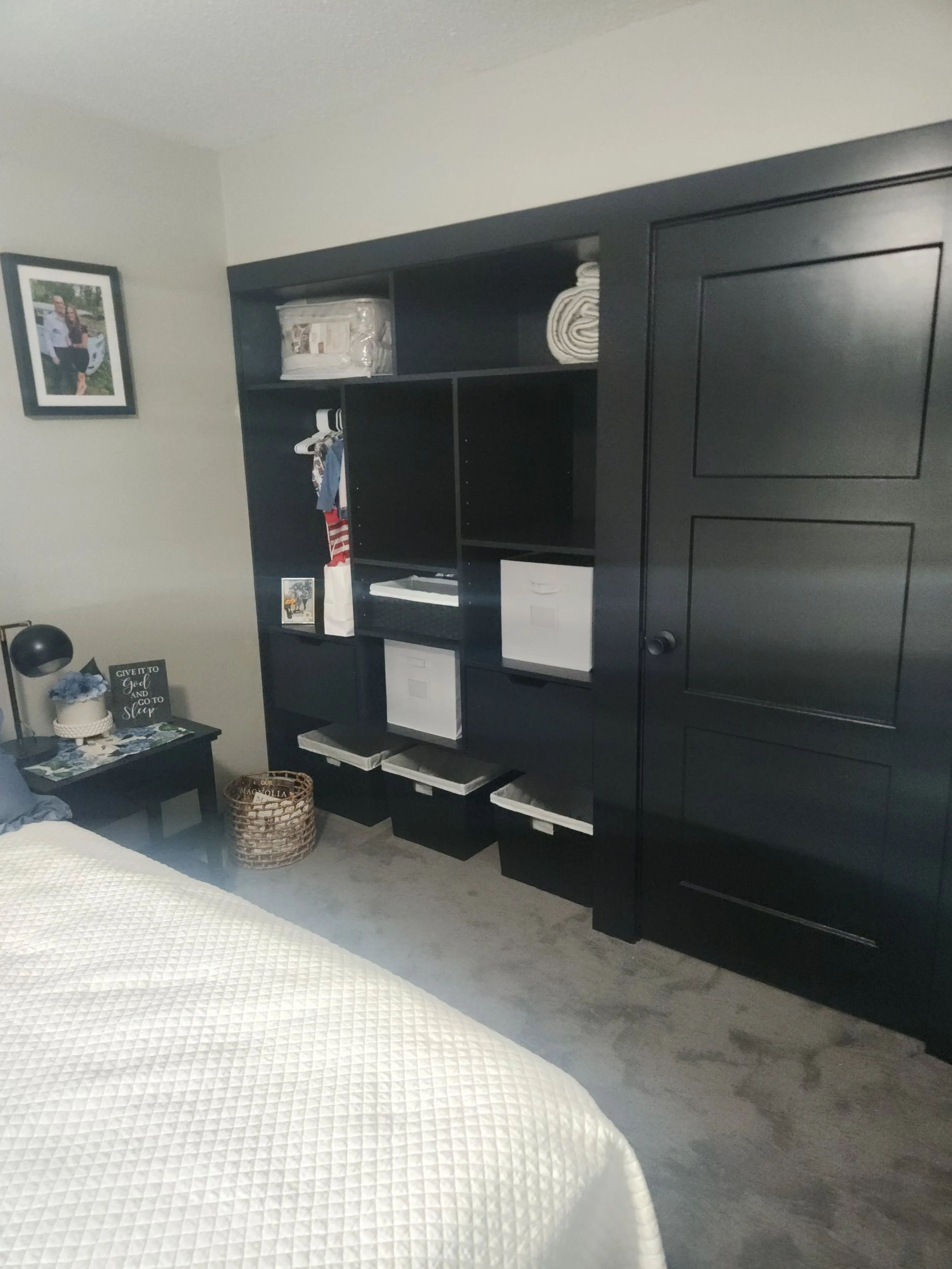 A bedroom with a black storage unit against the wall, containing folded linens and hanging clothes, a side table with a lamp and decorative items, a framed picture on the wall, and part of a bed with a white quilt.