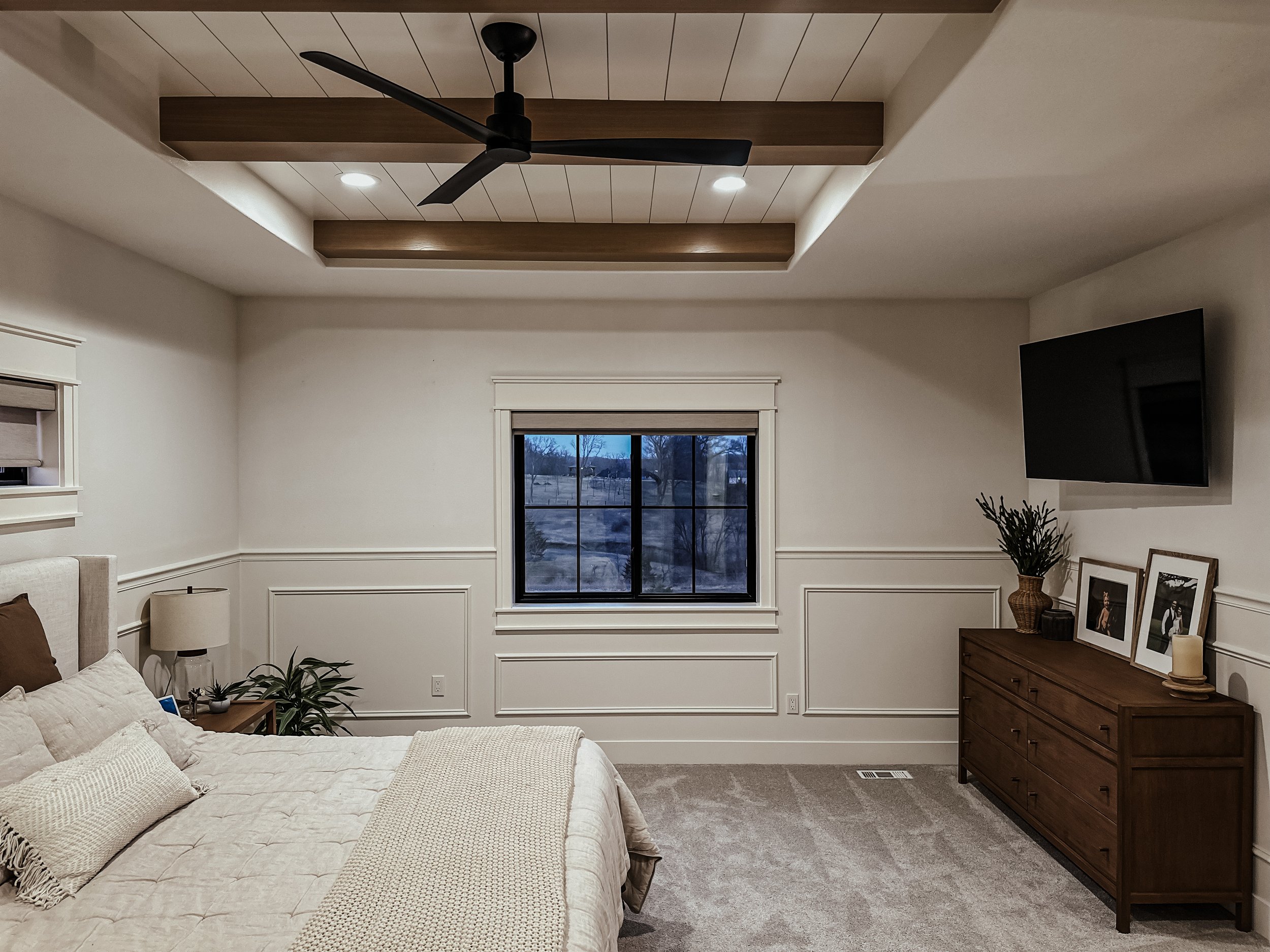 A bedroom with a beige bed, a nightstand with a lamp and plant, a window showing an outdoor view, a wooden dresser with photo frames, plant, and candle, a mounted TV, and a ceiling fan.