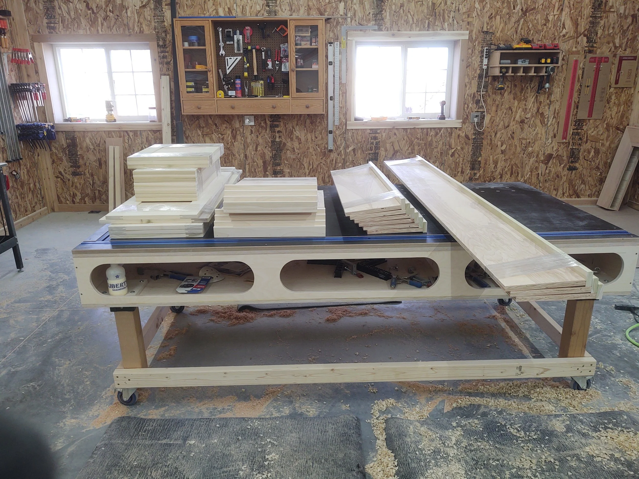 Inside a woodworking workshop with a large workbench holding woods and tools, and shelves and windows on plywood walls.