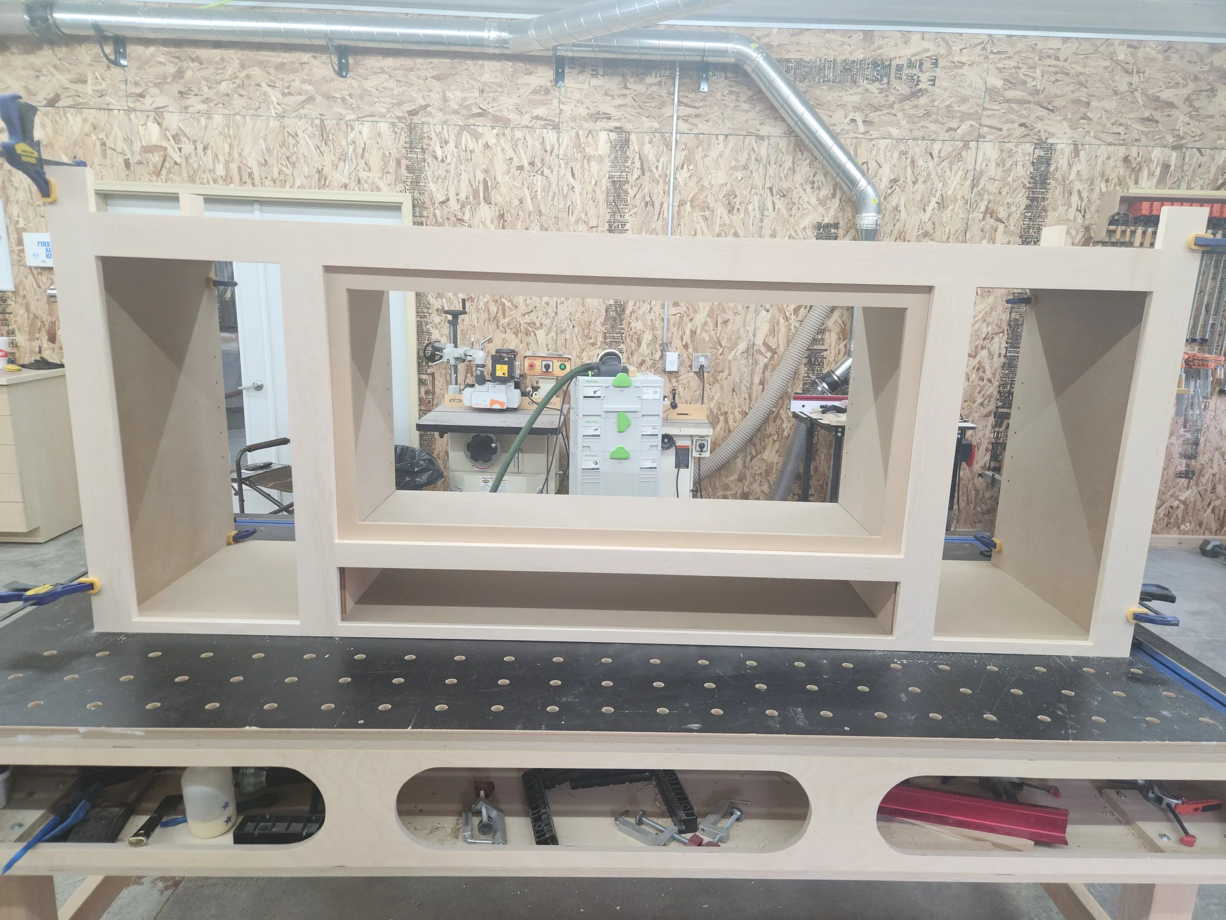 A wooden workbench in a workshop with a large window frame made of wood resting on it, various tools and equipment around, and a pegboard wall with clamps and supplies.