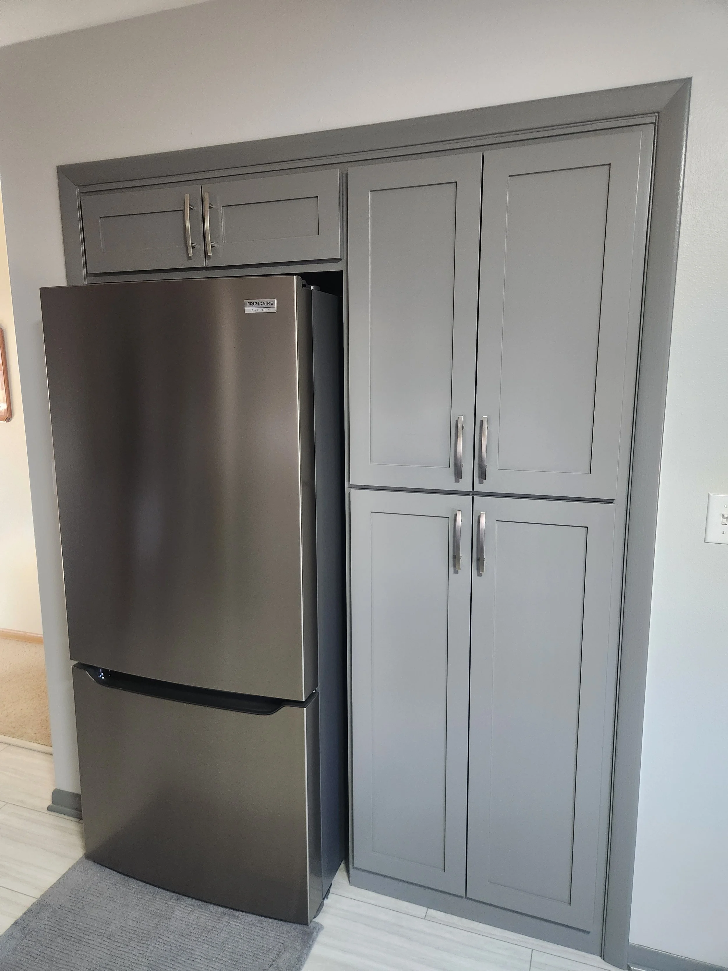 Stainless steel refrigerator next to gray kitchen cabinets with handles, located in a kitchen with light-colored flooring and a small gray rug.