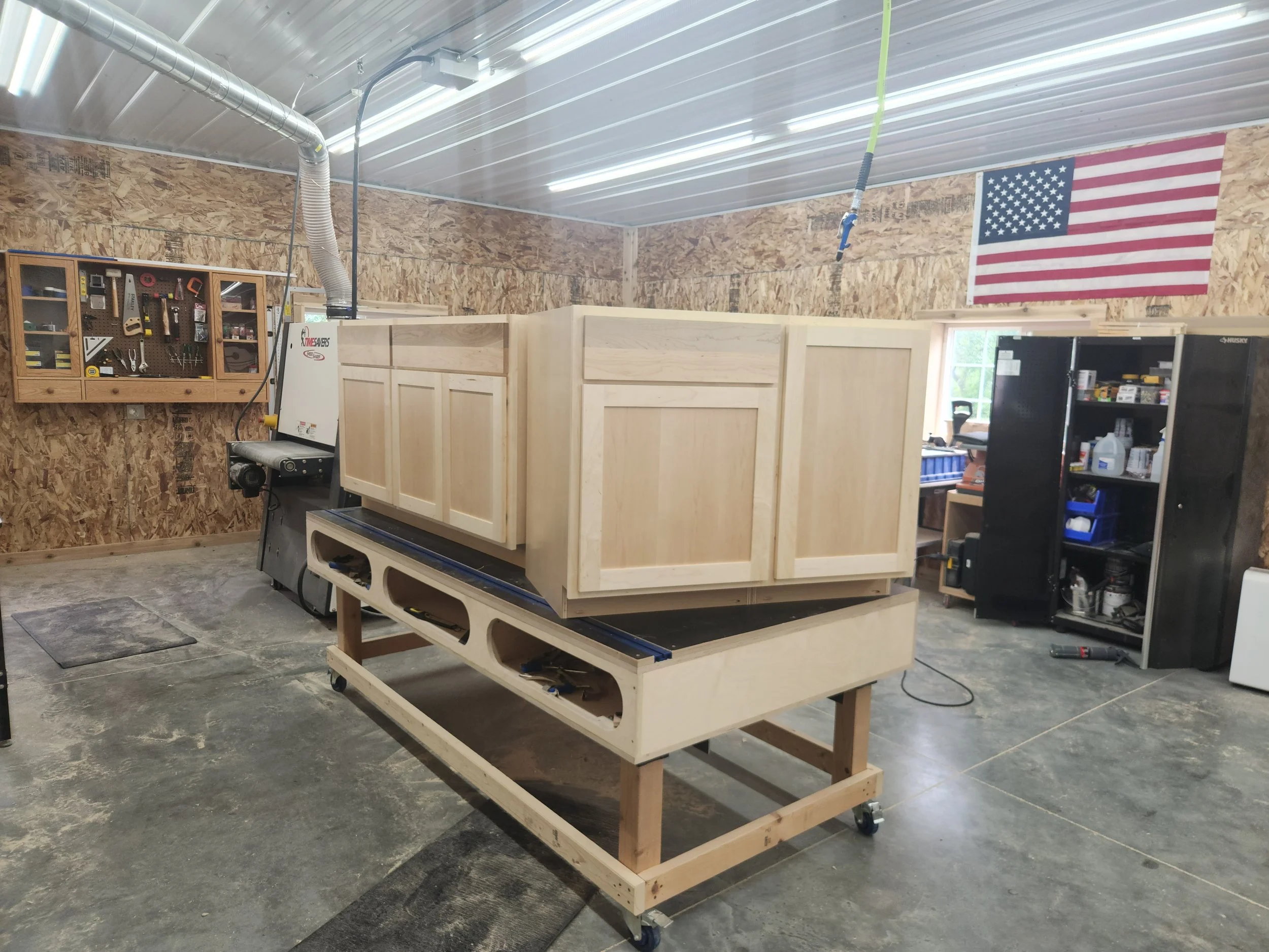 Workshop with woodwork station, large wooden cabinet on a workbench, American flag on the wall, pegboard with tools, workshop tools and supplies on shelves.