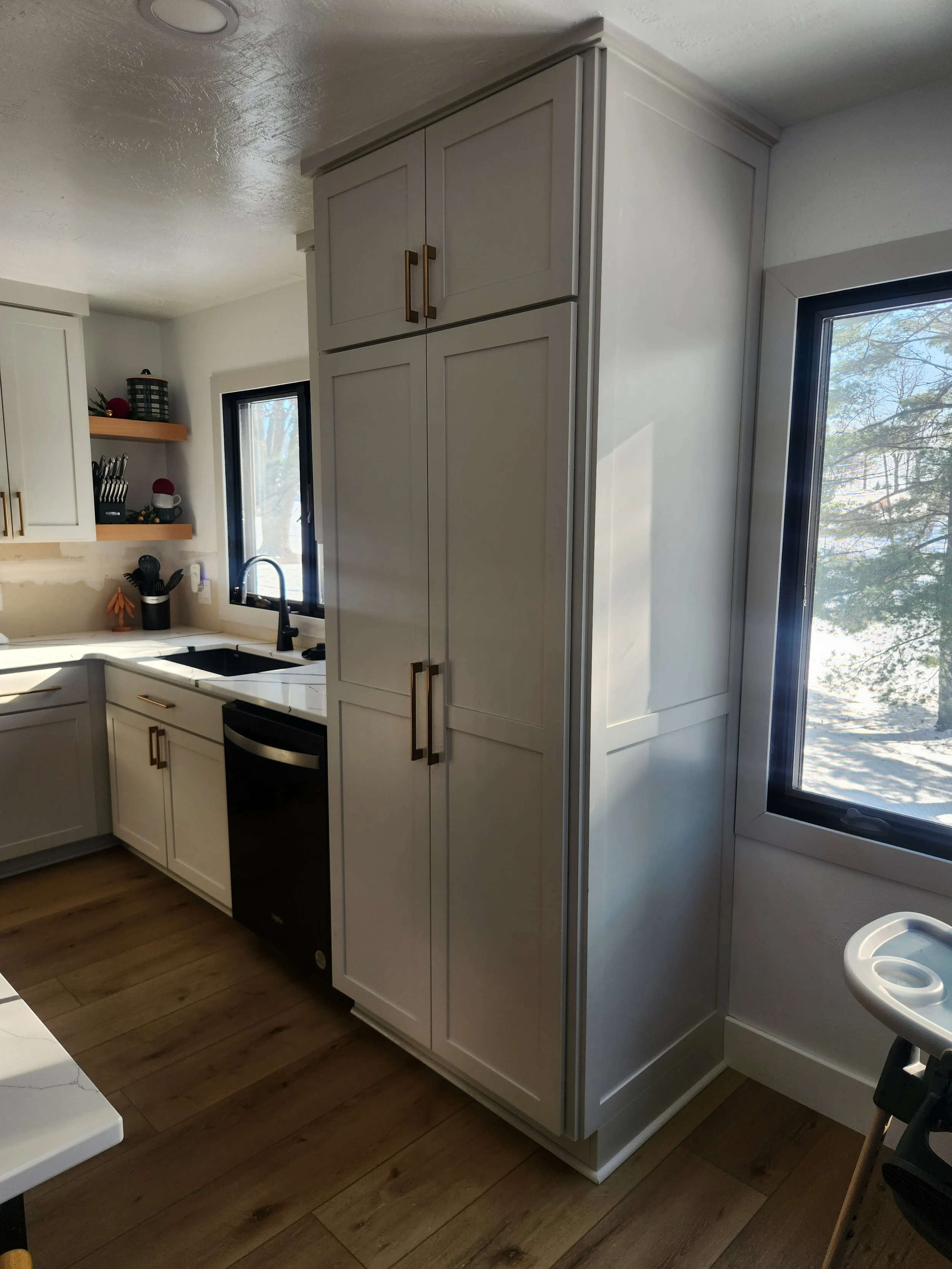 Kitchen with white cabinets, a window above the sink, and a built-in refrigerator. Light wood flooring and a high chair are visible.