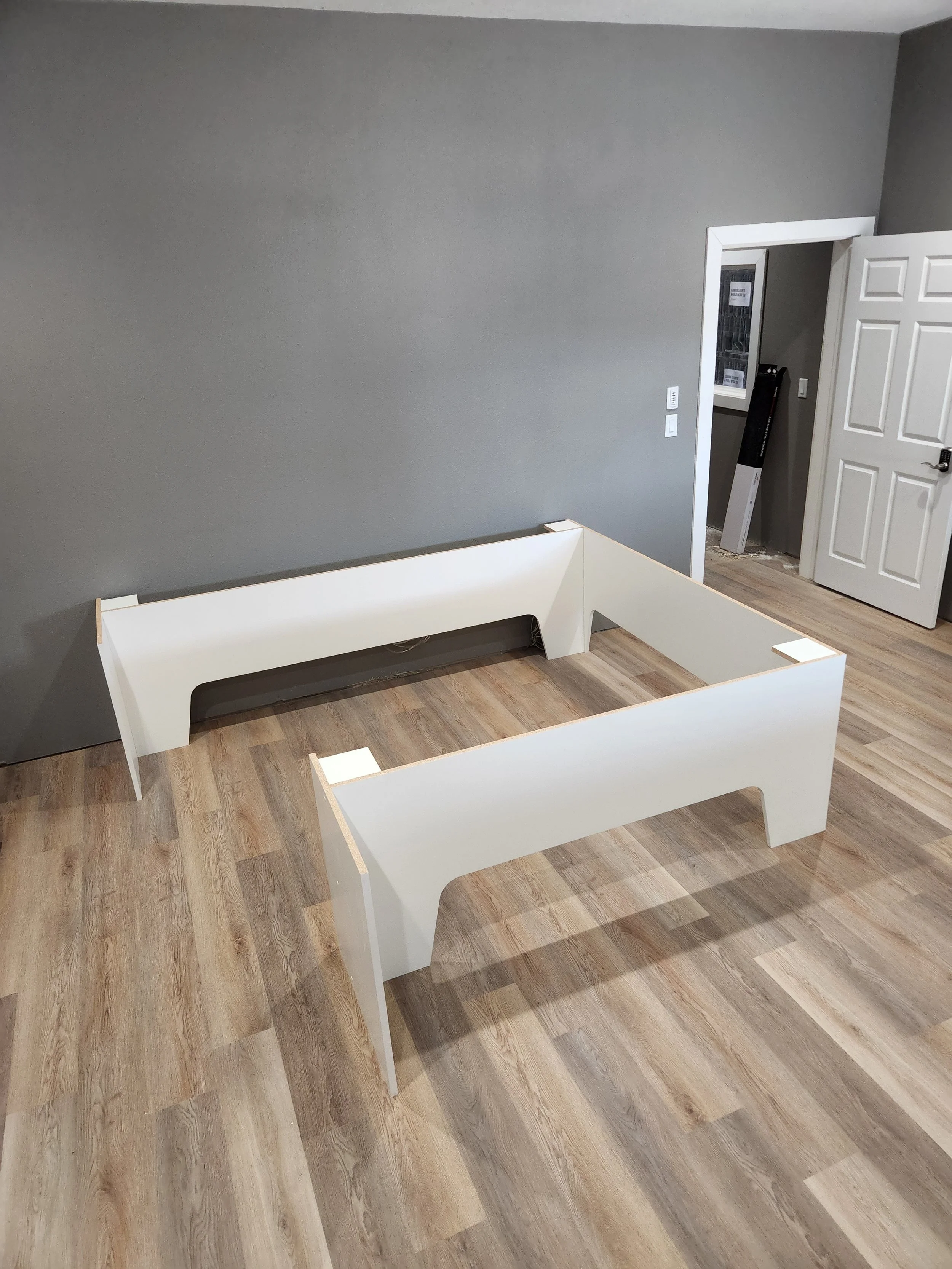 Unassembled white bed frame in a room with gray walls and wooden flooring.