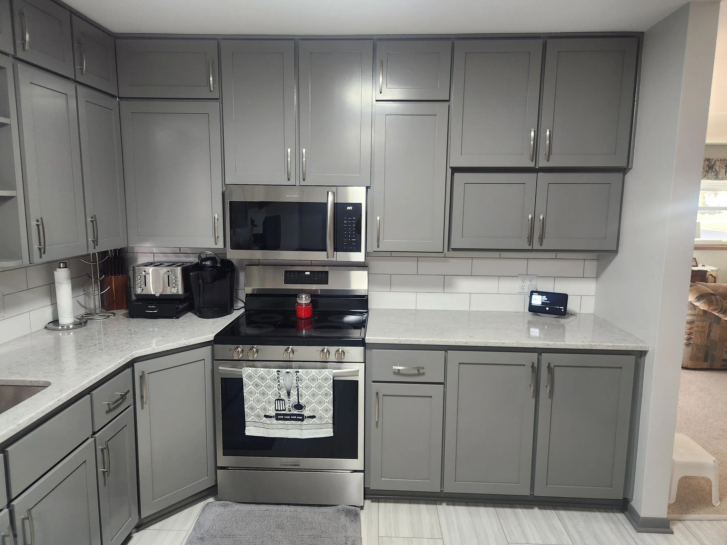Grey kitchen cabinets with stainless steel appliances, including a microwave and oven, on a white countertop with a black coffee machine, toaster, and small tablet.