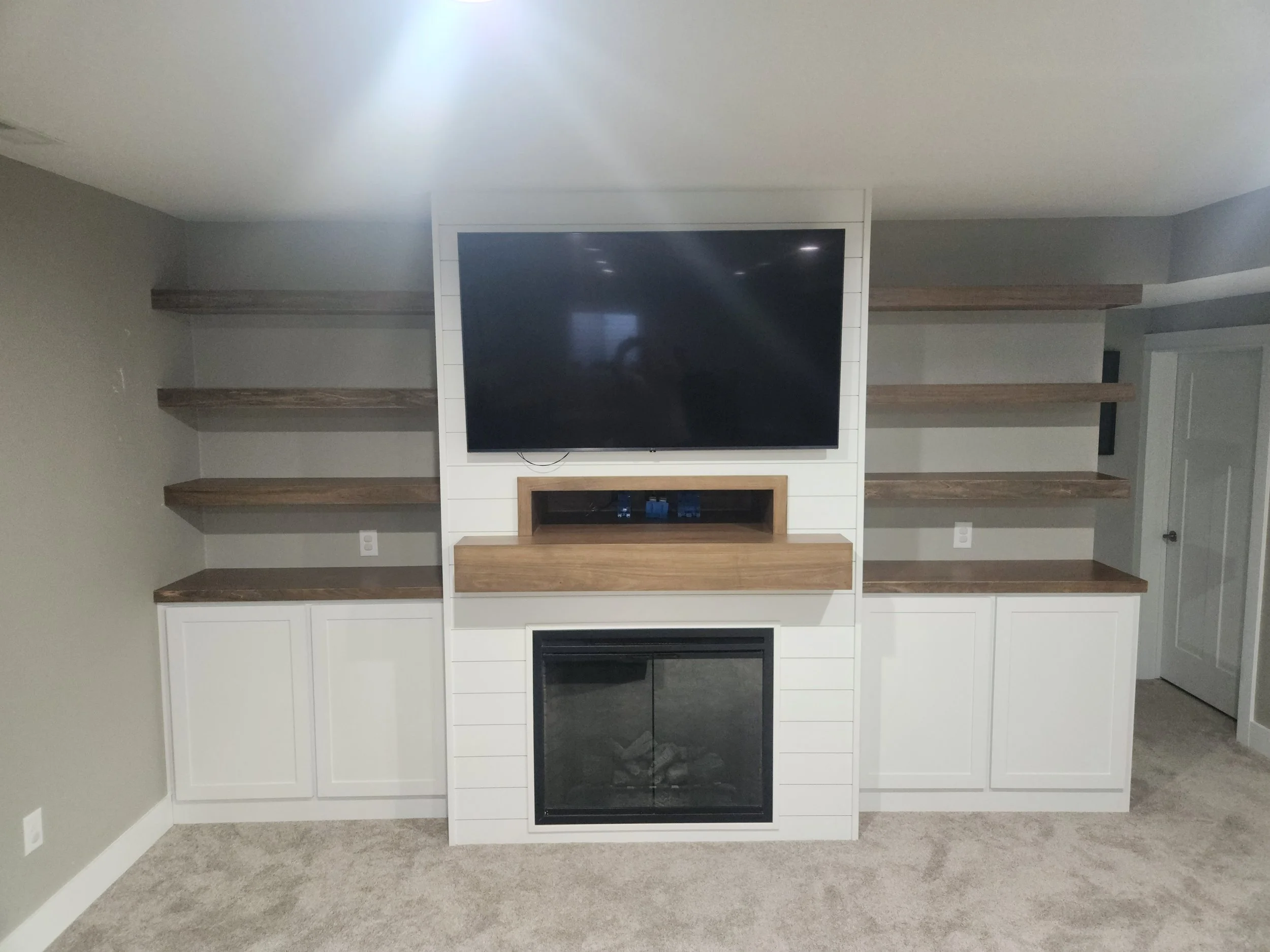 Living room entertainment center with a large TV mounted above a fireplace, surrounded by wooden shelves and white cabinetry.