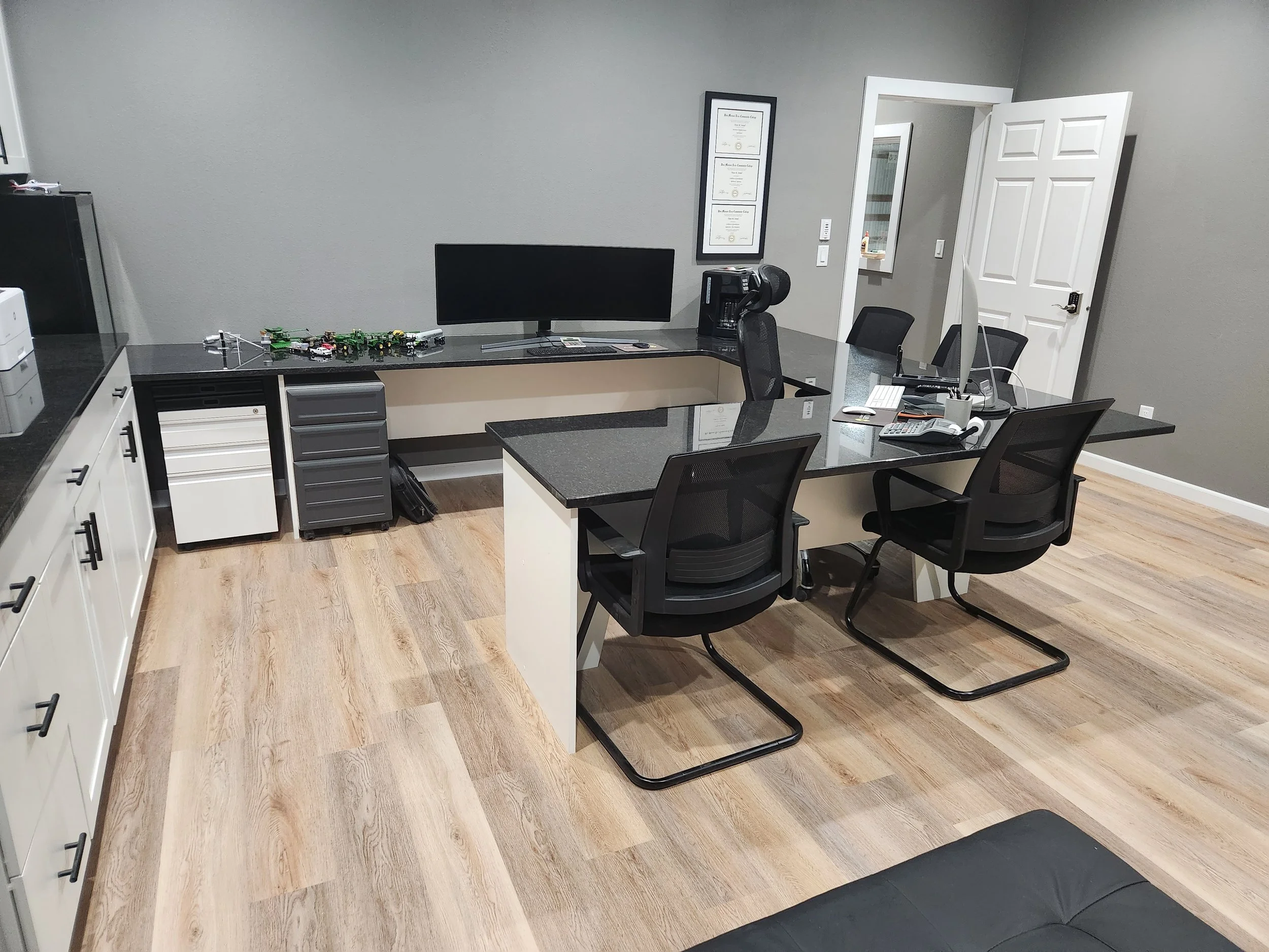 Office room with a black desk, three black chairs, a computer, monitor, desk phone, and office supplies. Gray walls, wood floor, and framed certificates on the wall.