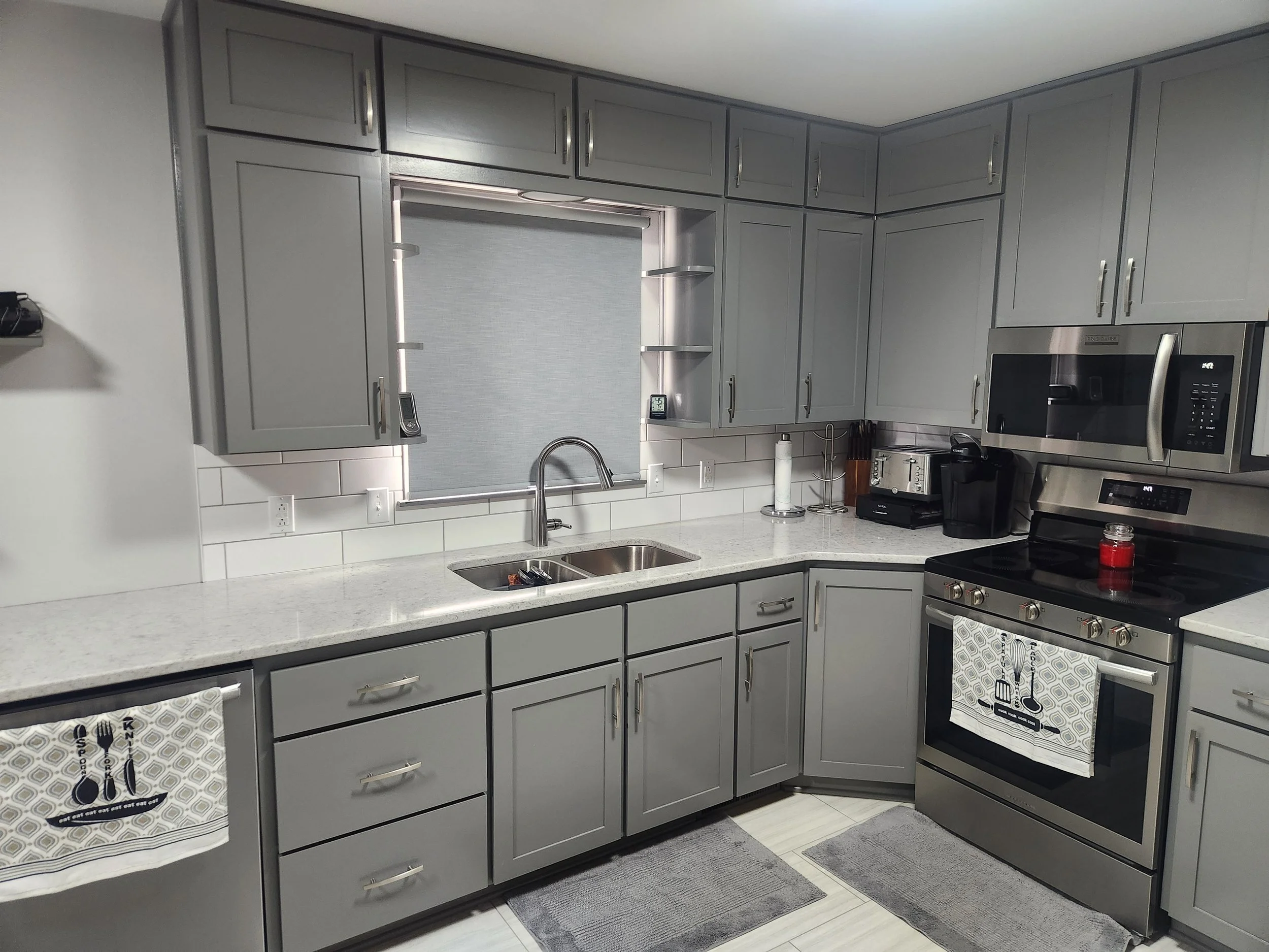 Gray kitchen cabinets with white speckled countertops, stainless steel appliances including a stove, microwave, and coffee maker, a window with a gray shade, and various kitchen tools on the counter.