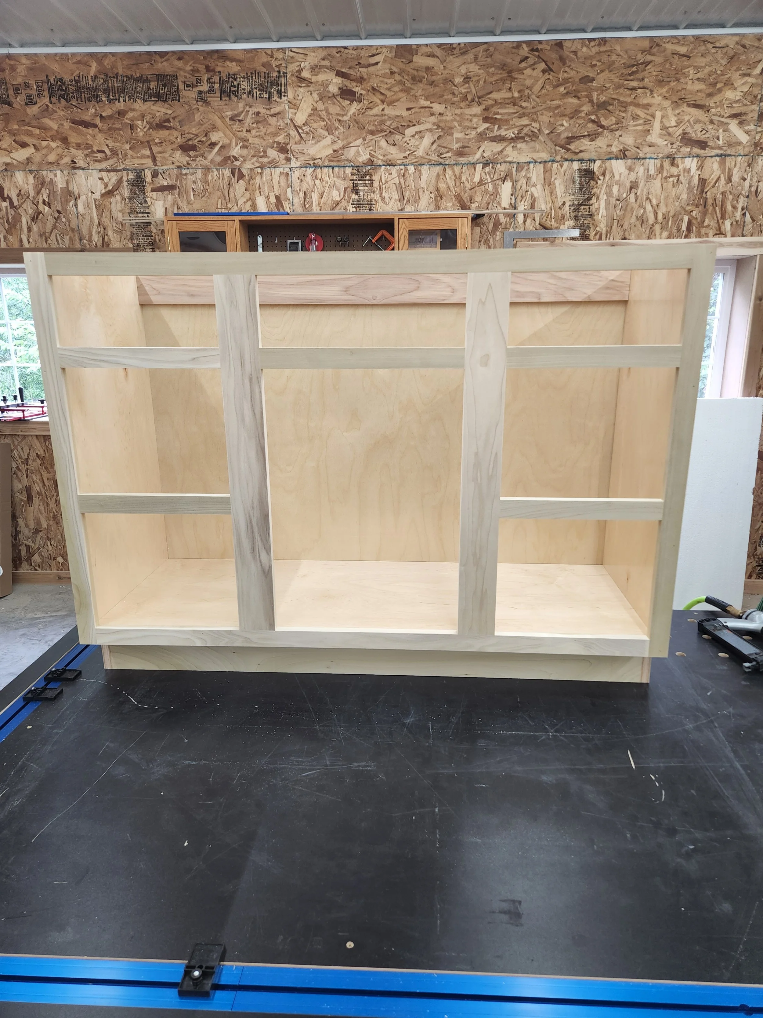Unfinished wooden shelf or frame being built in a workshop with tools on the work surface and a plywood wall in the background.