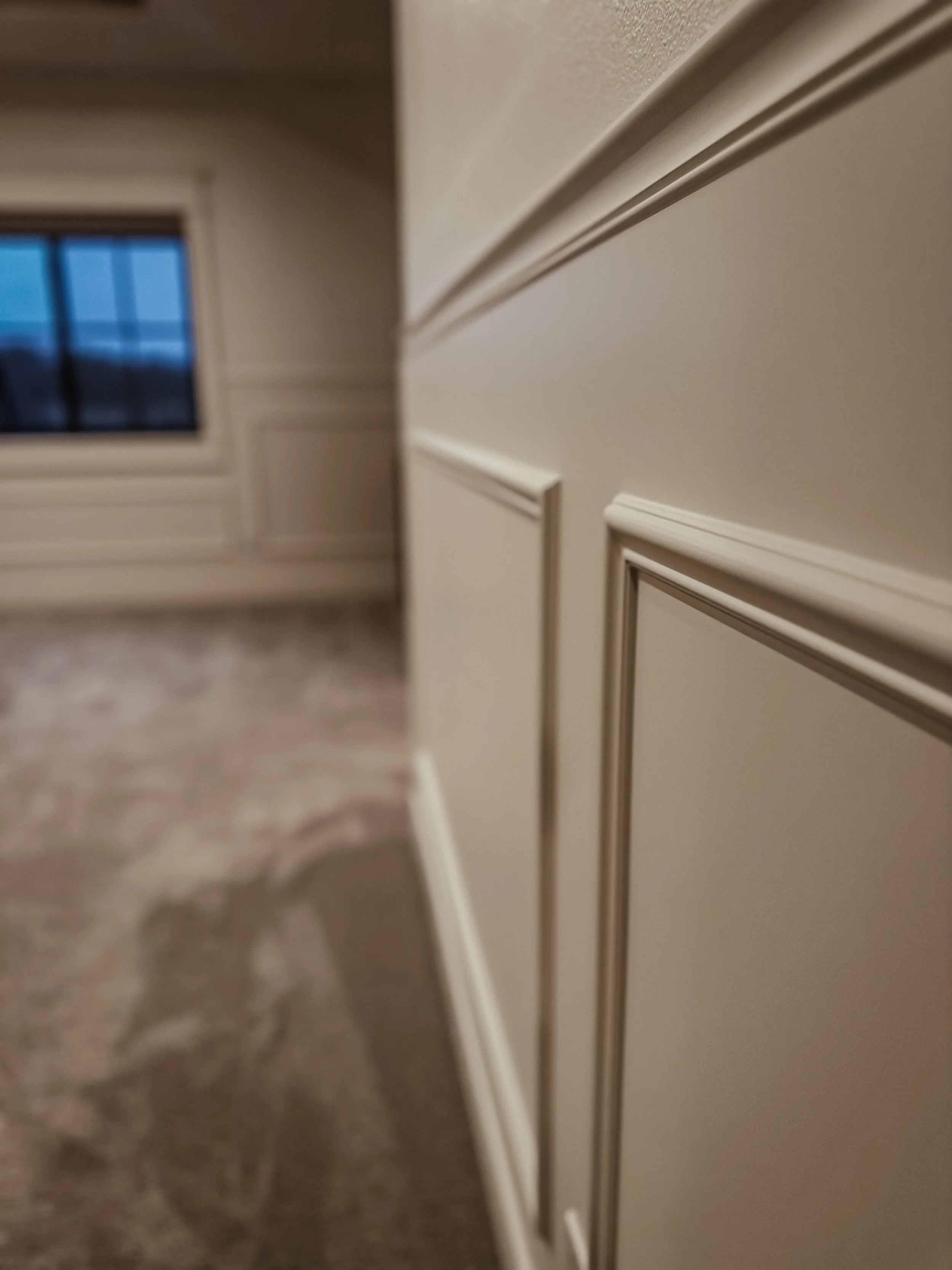 Close-up of cream-colored wainscoting or paneling on a wall, with a window visible in the background.