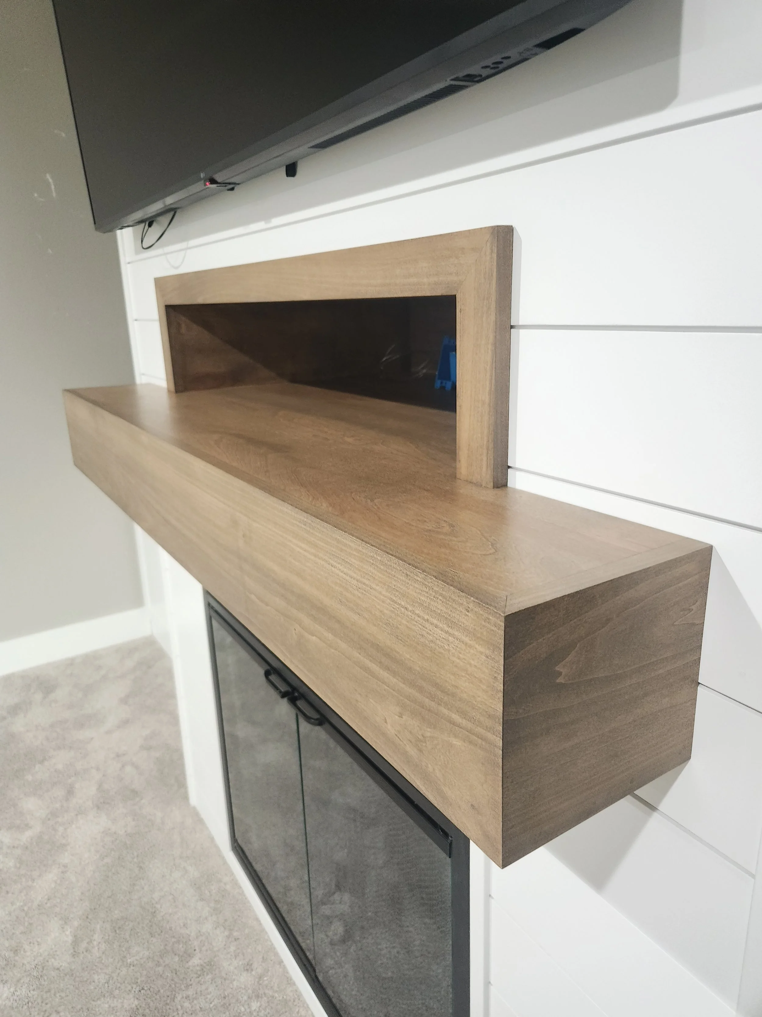 Wooden shelf mounted on a white wall above a black appliance with sliding doors, with a flat-screen TV above.
