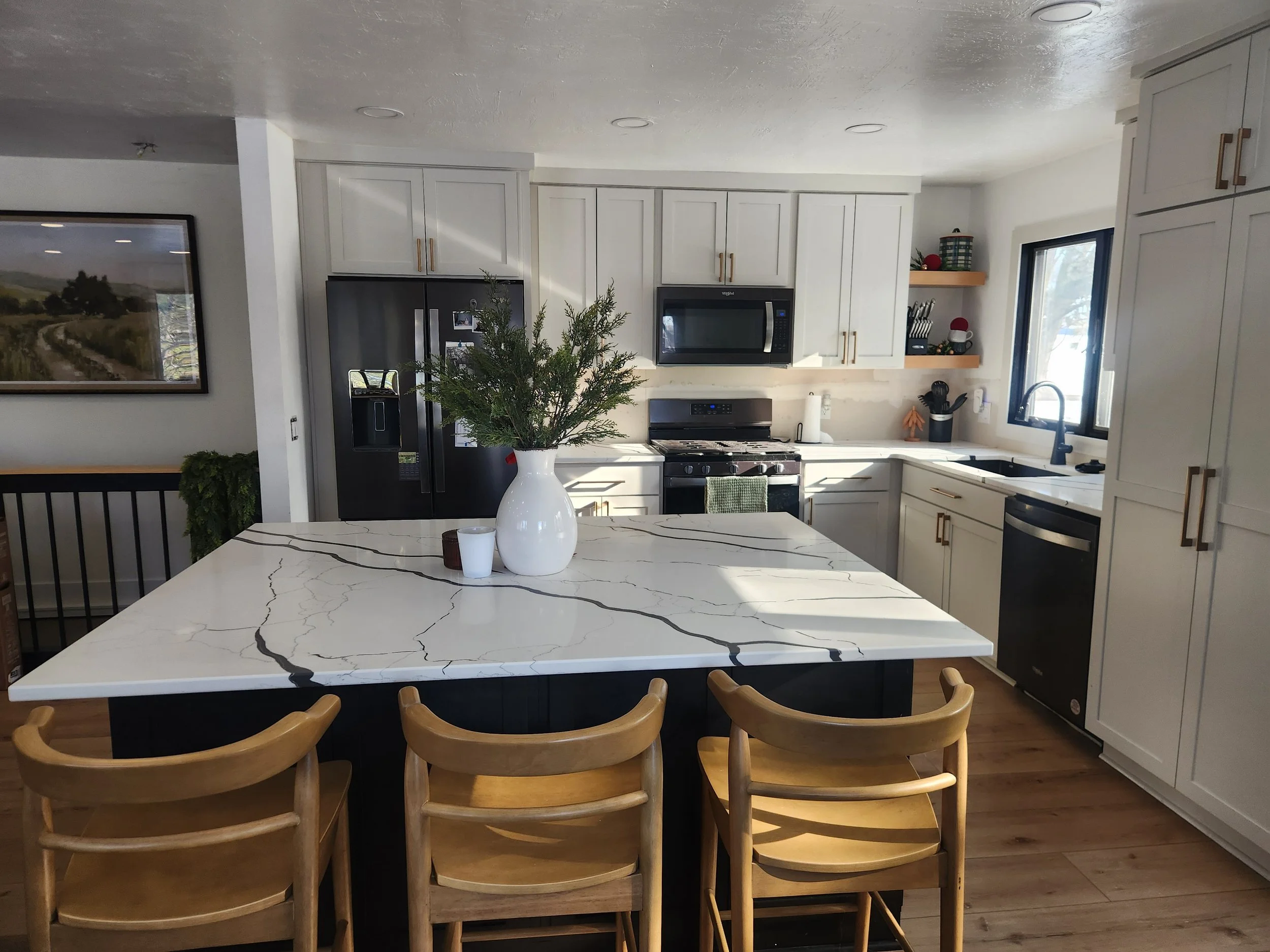 Modern kitchen with white cabinets, black appliances, and a large kitchen island with a white marble top. Four wooden chairs are around the island, and a white vase with greenery, a small candle, and a box are on top. There is a window with natural l