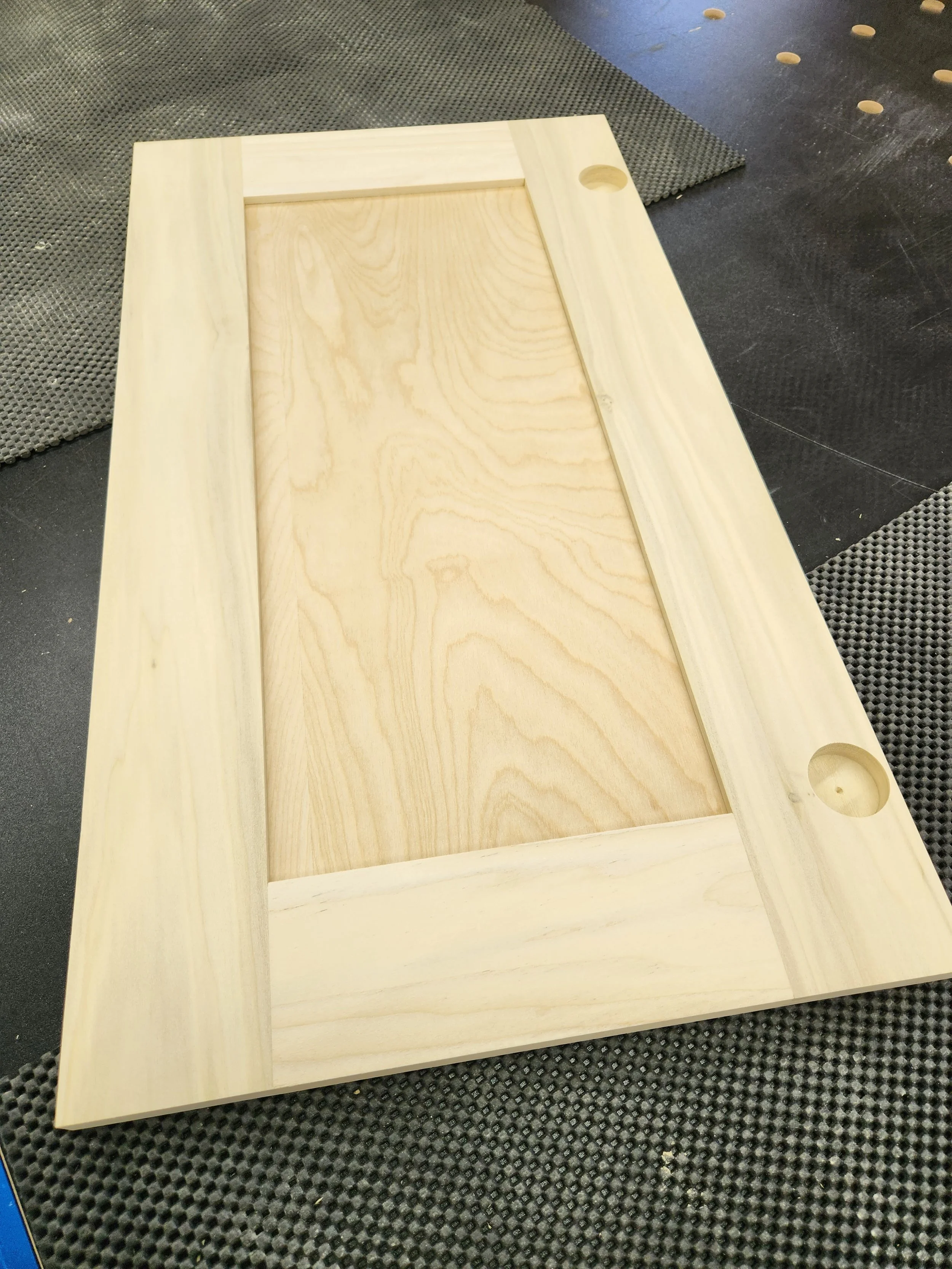 Wooden door panel with two circular holes, possibly for door handles, lying on a workbench.