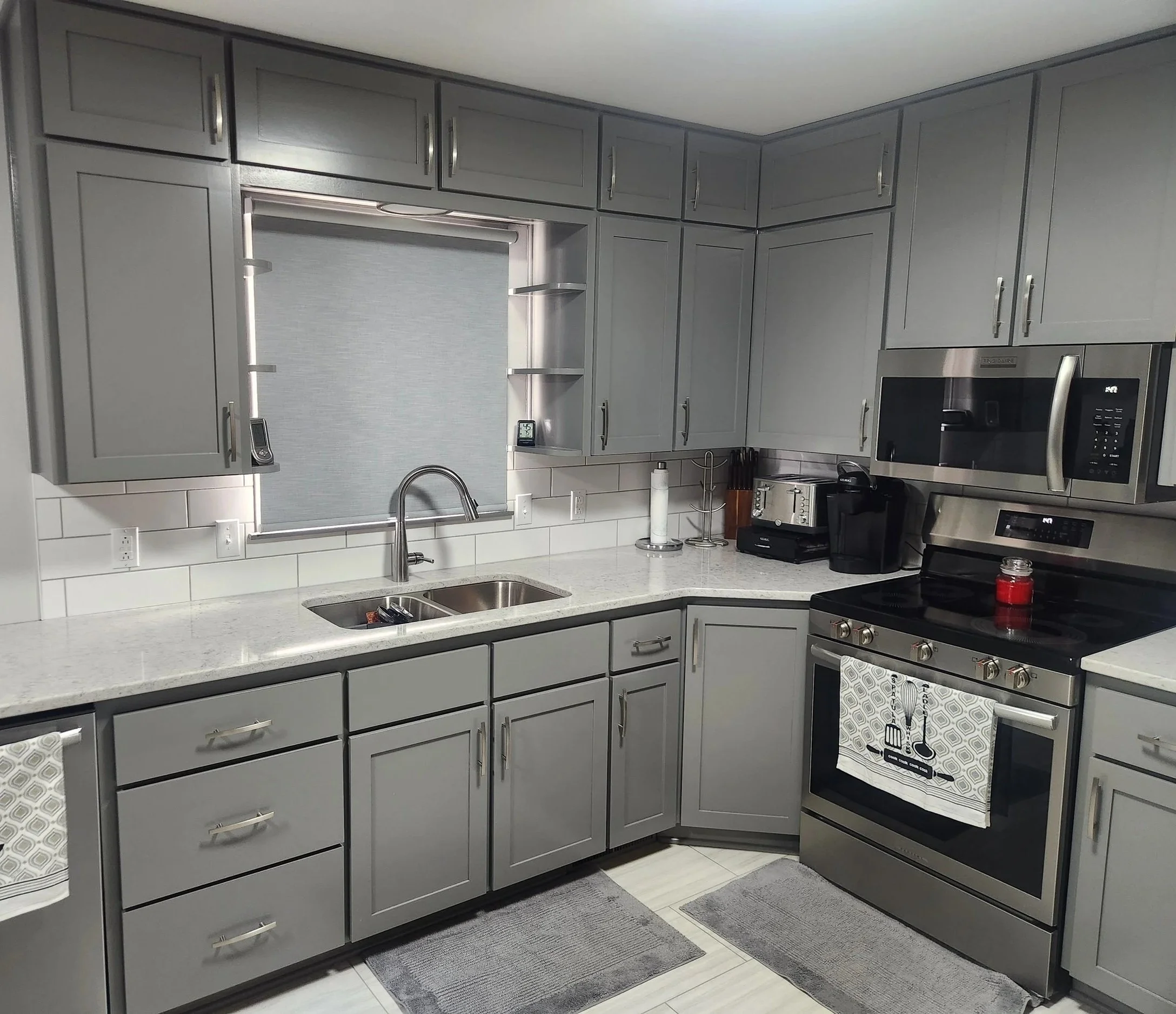 Grey kitchen with stainless steel appliances, including microwave, oven, and toaster, white countertop, grey cabinets, window with grey shade, and various kitchen utensils and appliances on counter.