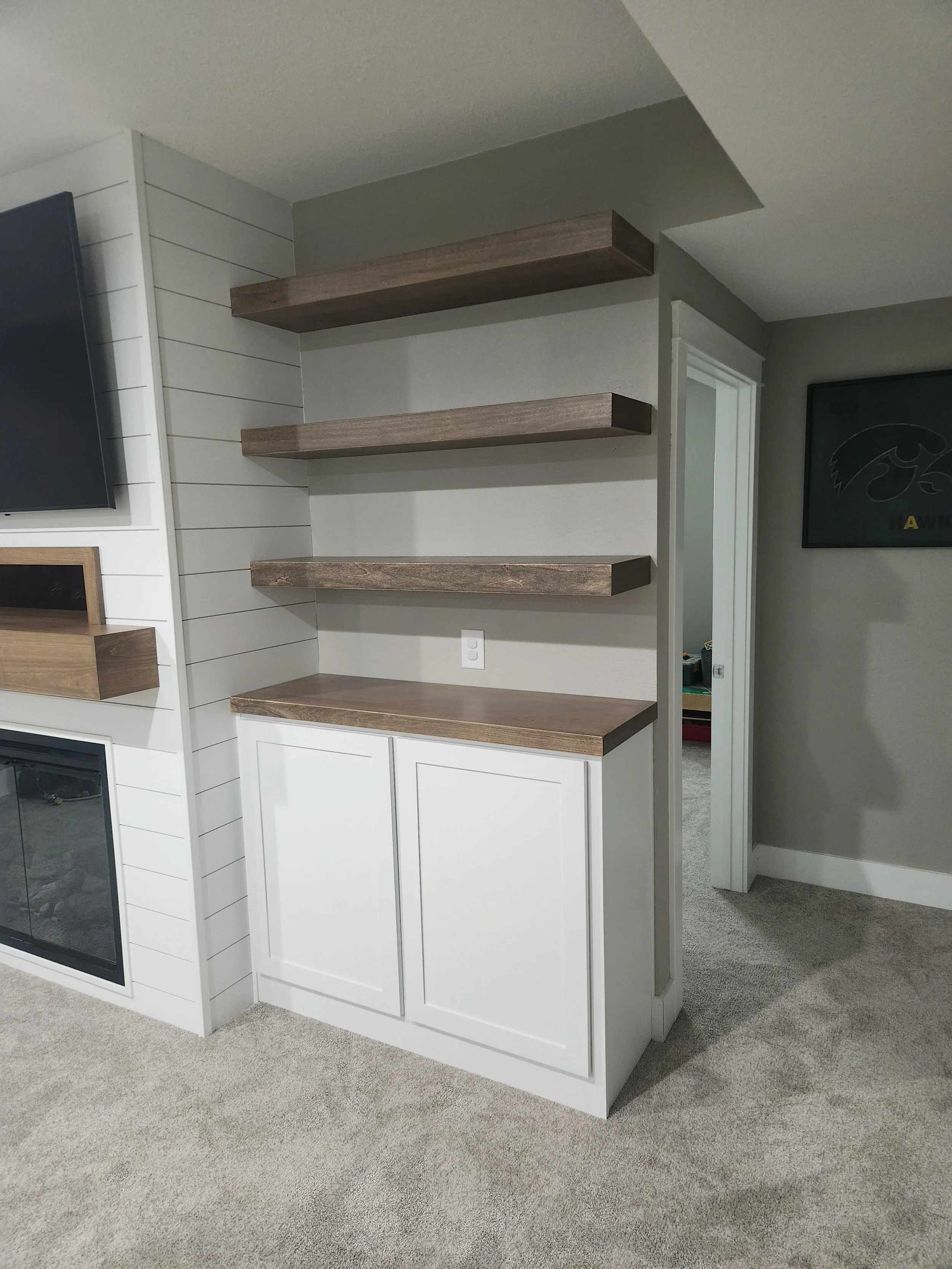 Living room with white built-in cabinet, three wooden shelves, a wall outlet, and a TV mounted on the wall. Carpeted floor and a doorway leading to another room.
