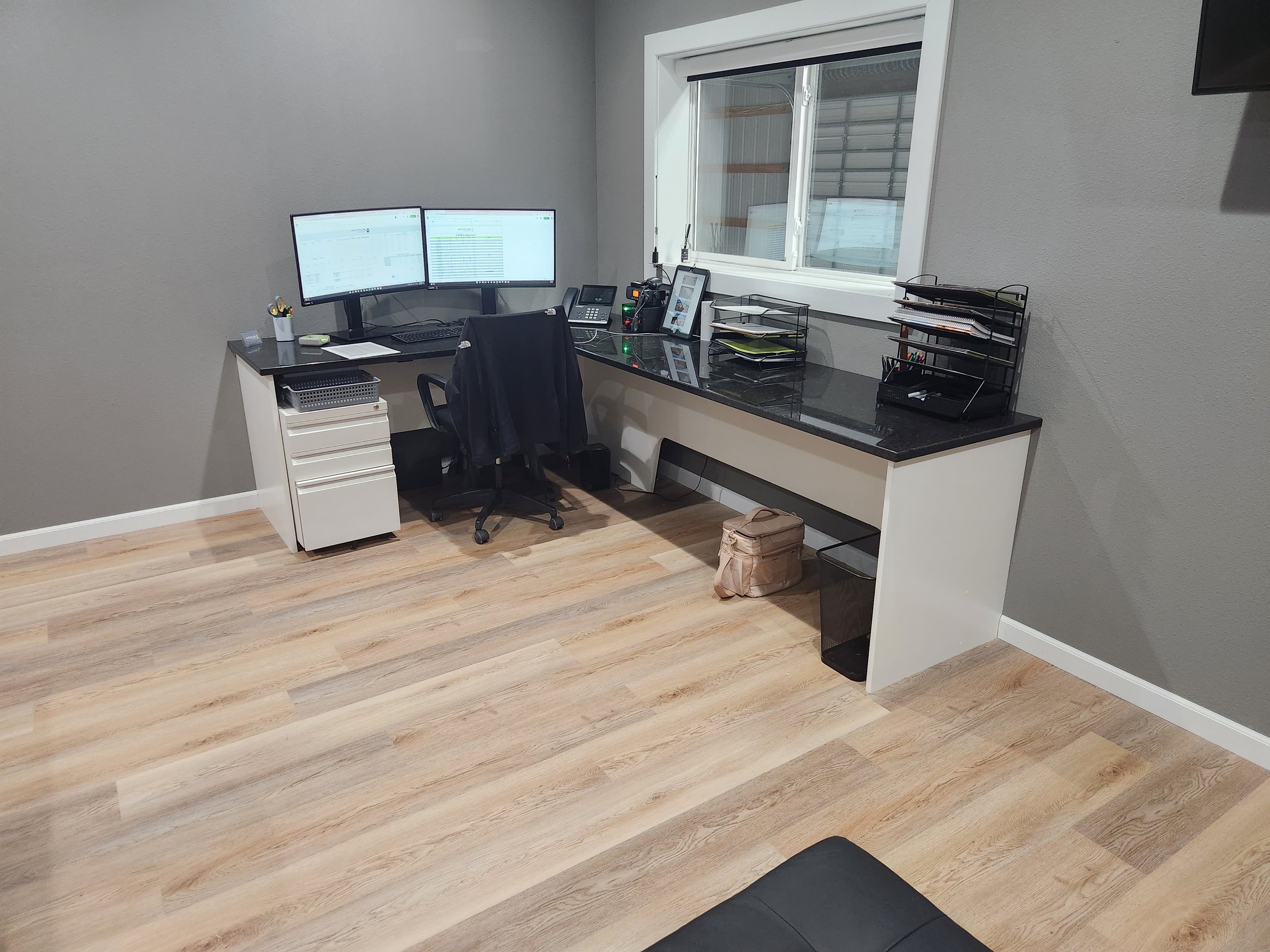 Empty office with a desk, three monitors, a black office chair, and various office supplies, with a window and light wood flooring.