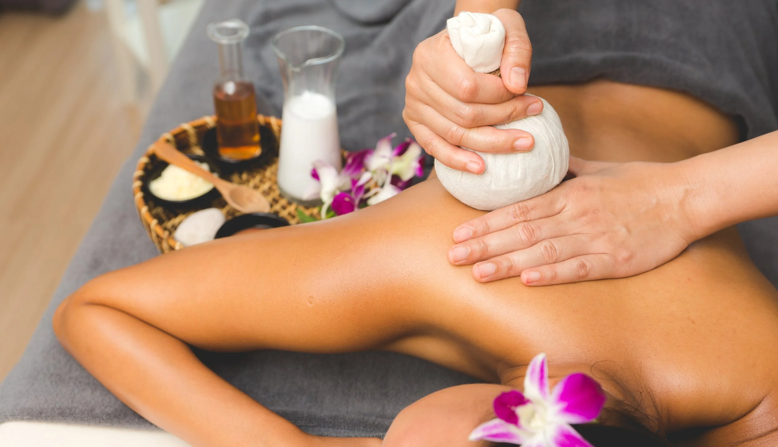 A person receiving a herbal compress massage on their lower back at a spa, with massage oils and flowers on a nearby tray.