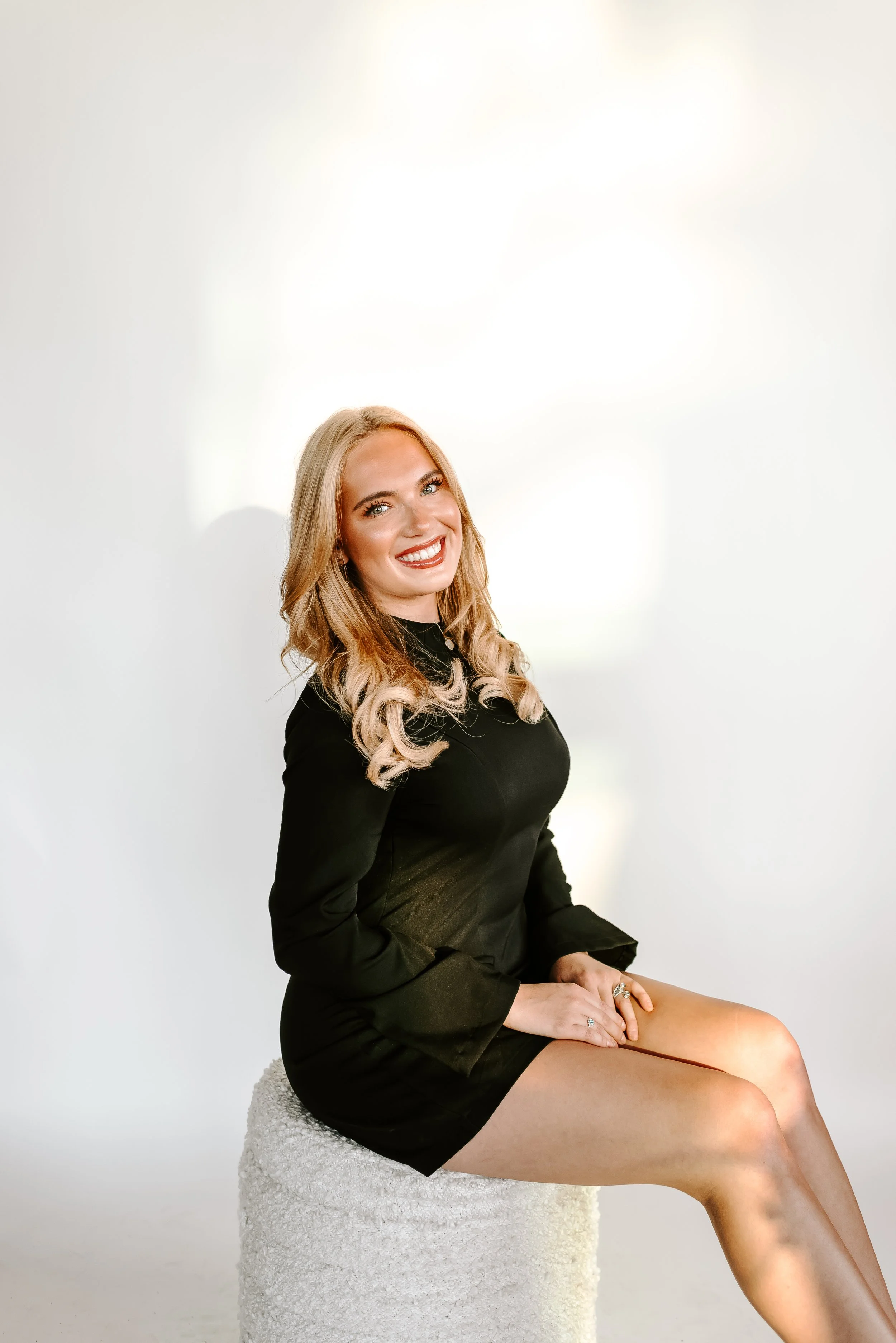 A smiling woman with blonde wavy hair in a black dress sitting on a white textured stool against a plain white background.