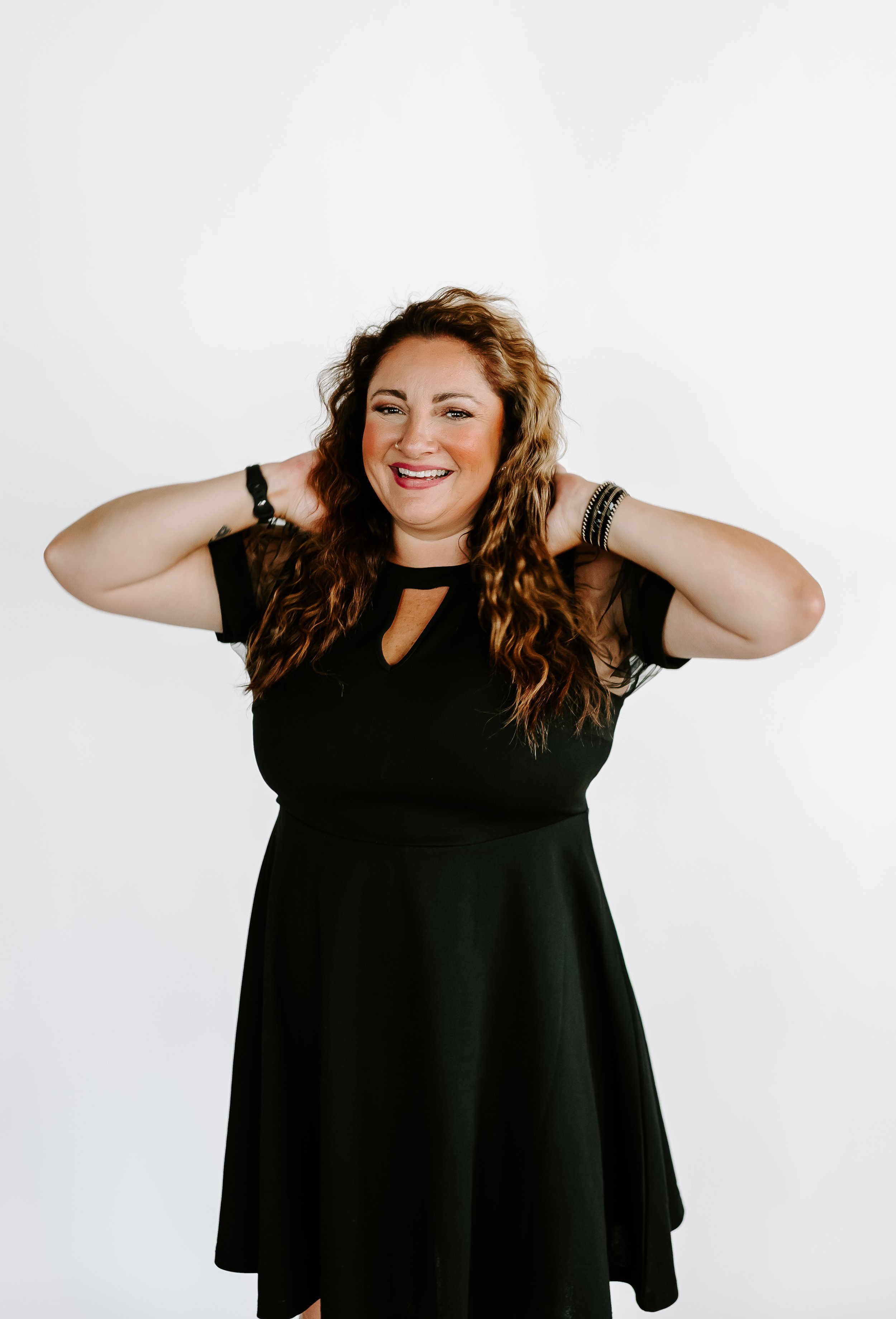 A woman with long, curly brown hair smiling, wearing a black dress, standing against a plain white background.