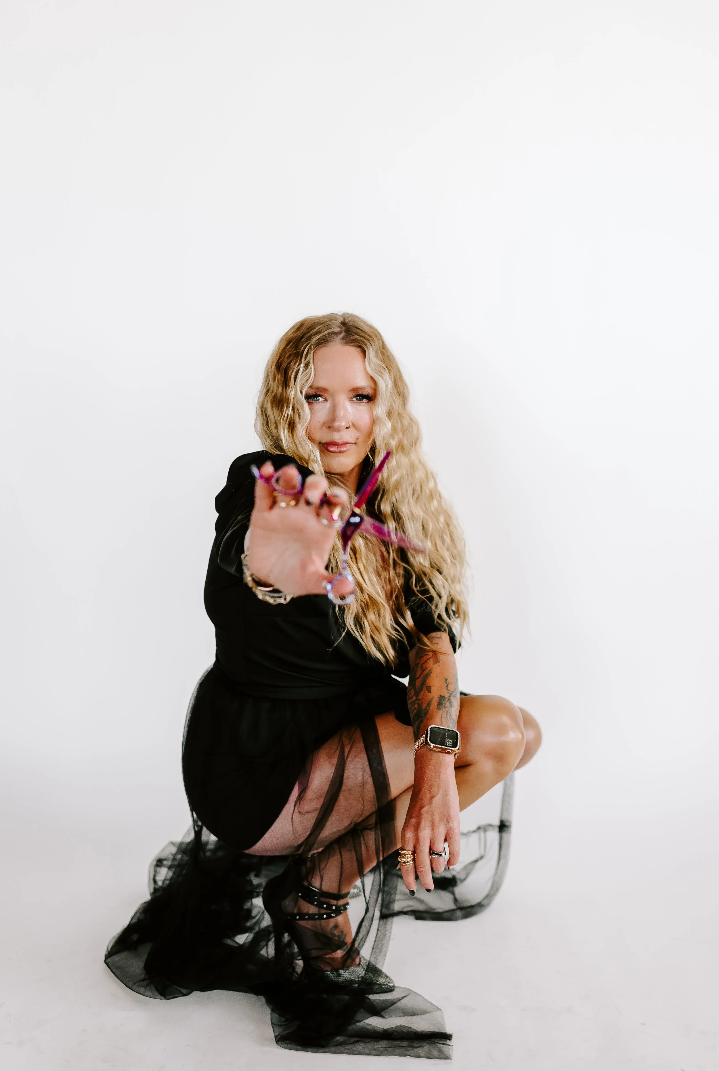 Woman with long blonde curly hair wearing black outfit and fishnet stockings squatting against white background, holding scissors towards the camera.